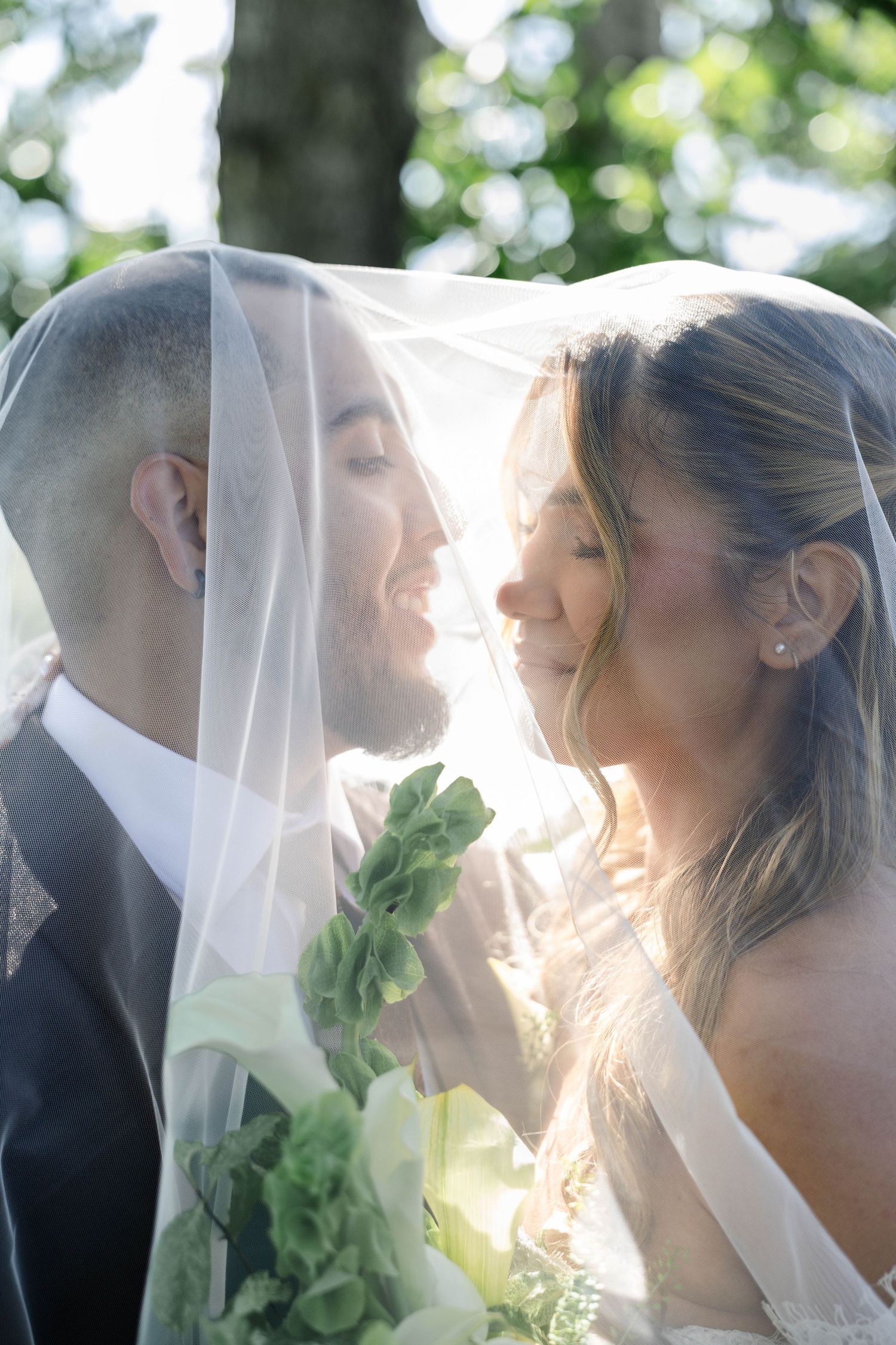 Bride and groom share a romantic moment under a sheer wedding veil outdoors, with soft sunlight and green trees.