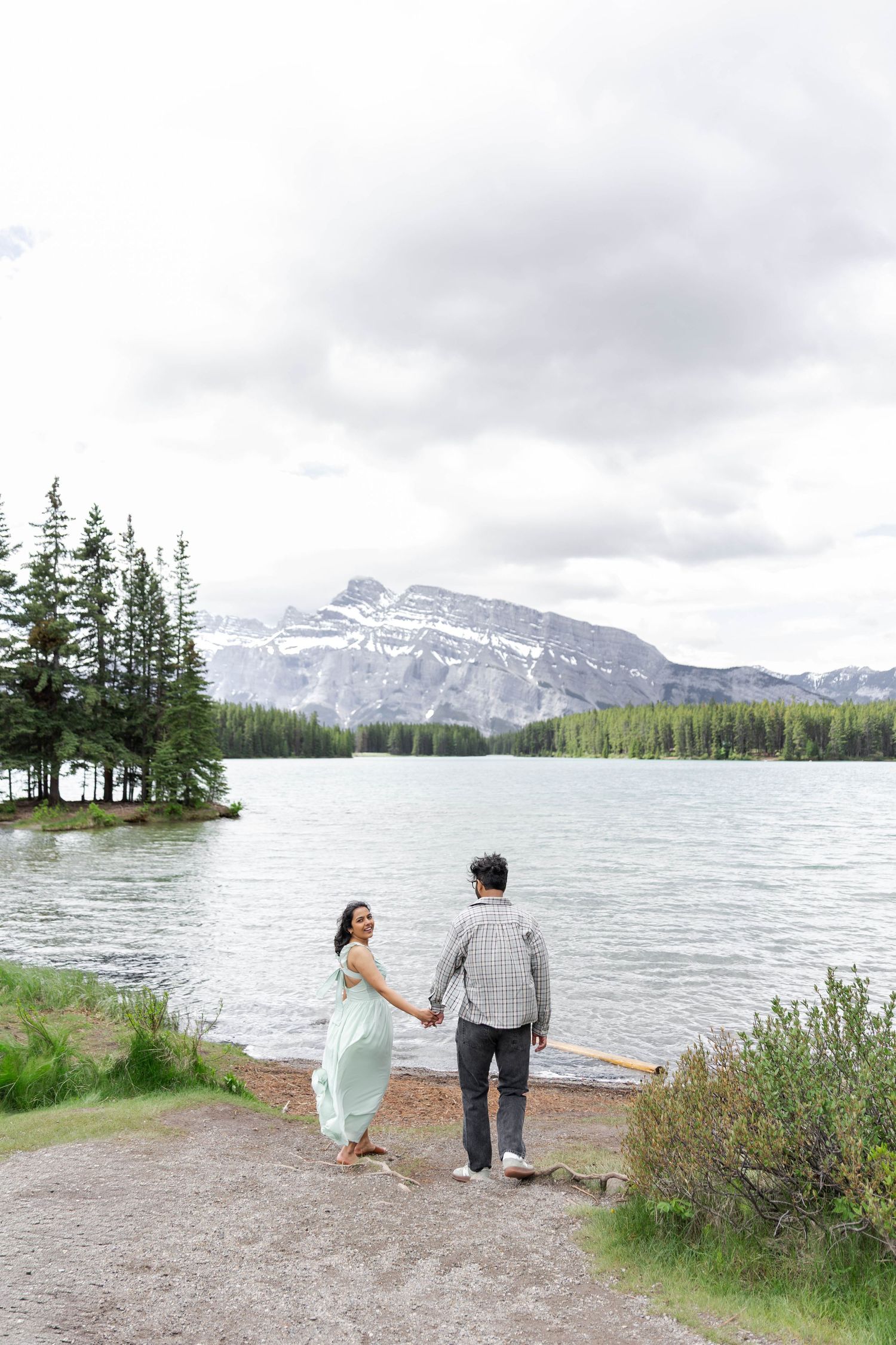 Couple holding hands by a mountain lake with snow-capped peaks and evergreen trees under a cloudy sky.