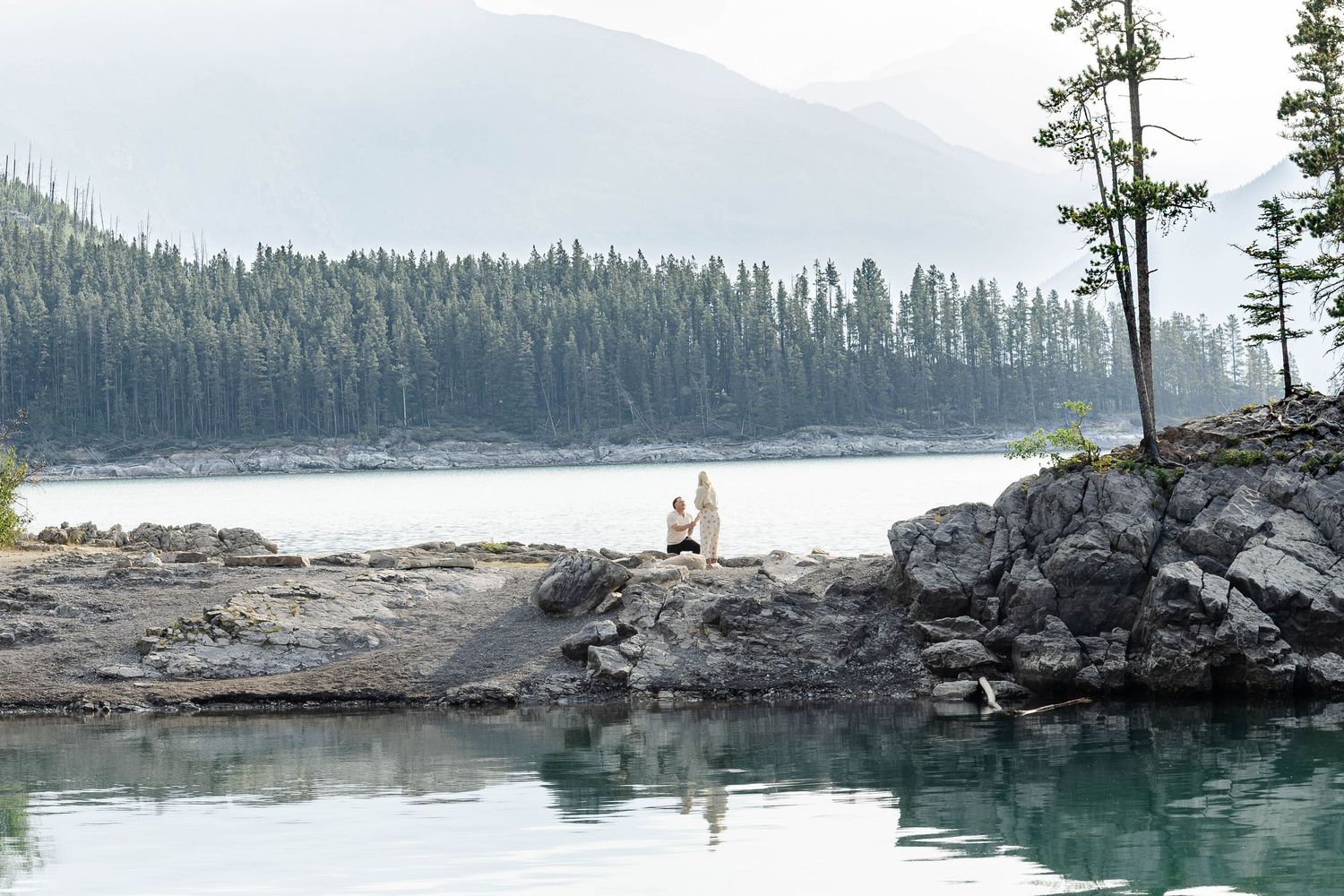 Two people sitting on rocky shoreline beside a tranquil turquoise lake surrounded by evergreen forest and misty mountains.Romantic couple