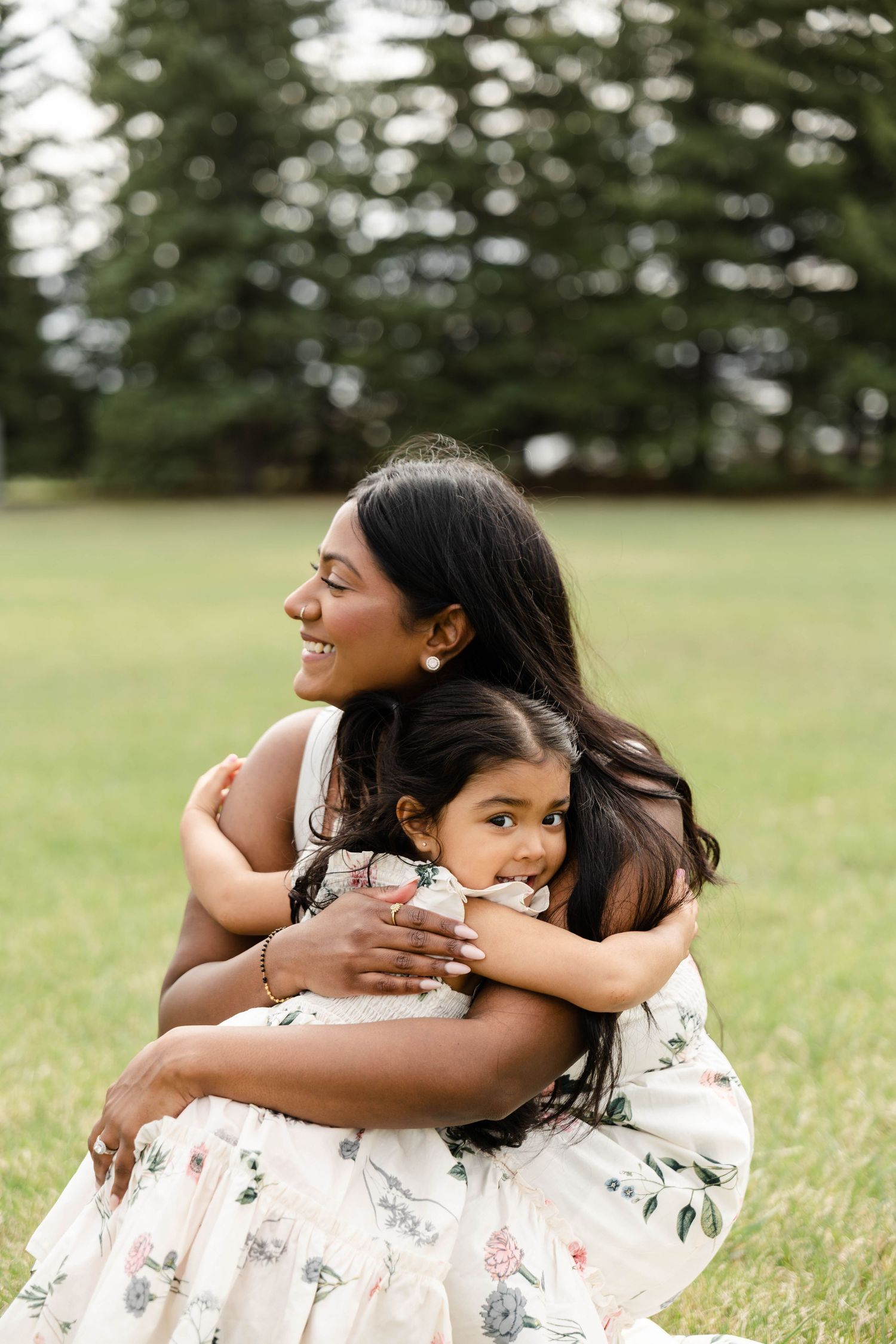 A smiling mother in a floral dress hugs two young daughters on a green lawn with pine trees in the background.