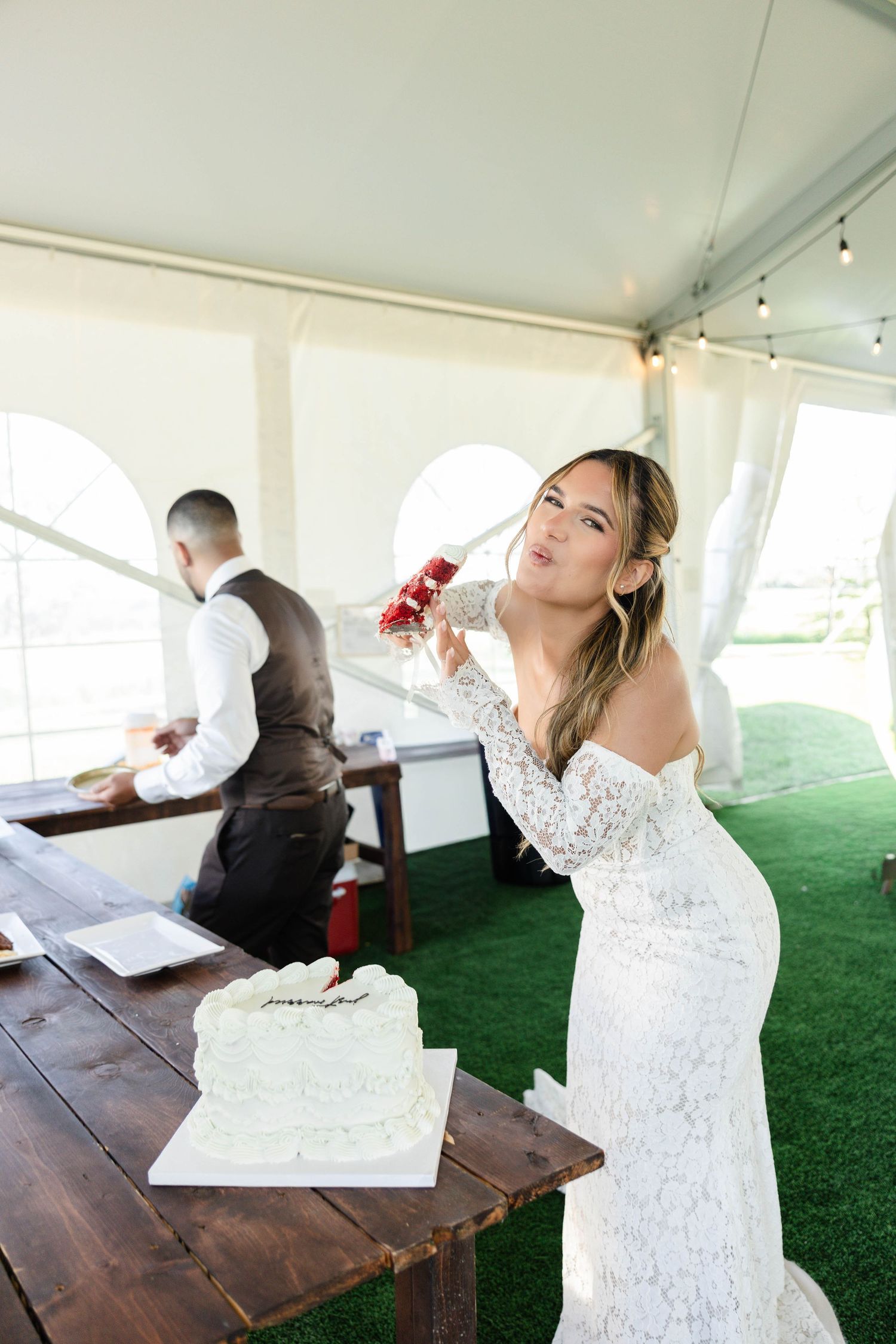 Bride in white lace gown poses with wedding cake under tent, holding red floral arrangement at outdoor reception.
