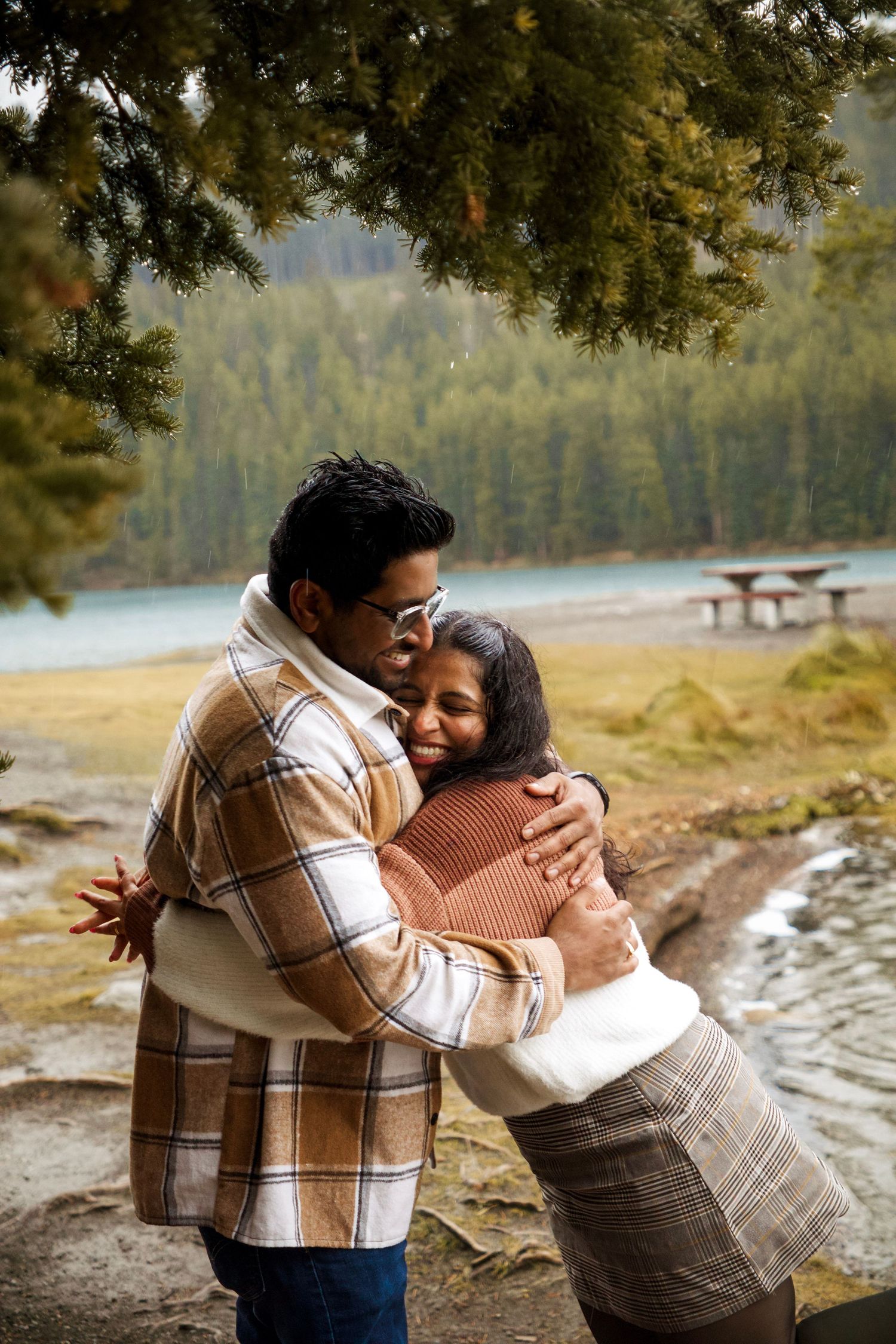 Couple embracing warmly near a turquoise lake surrounded by pine trees in an autumn forest setting.