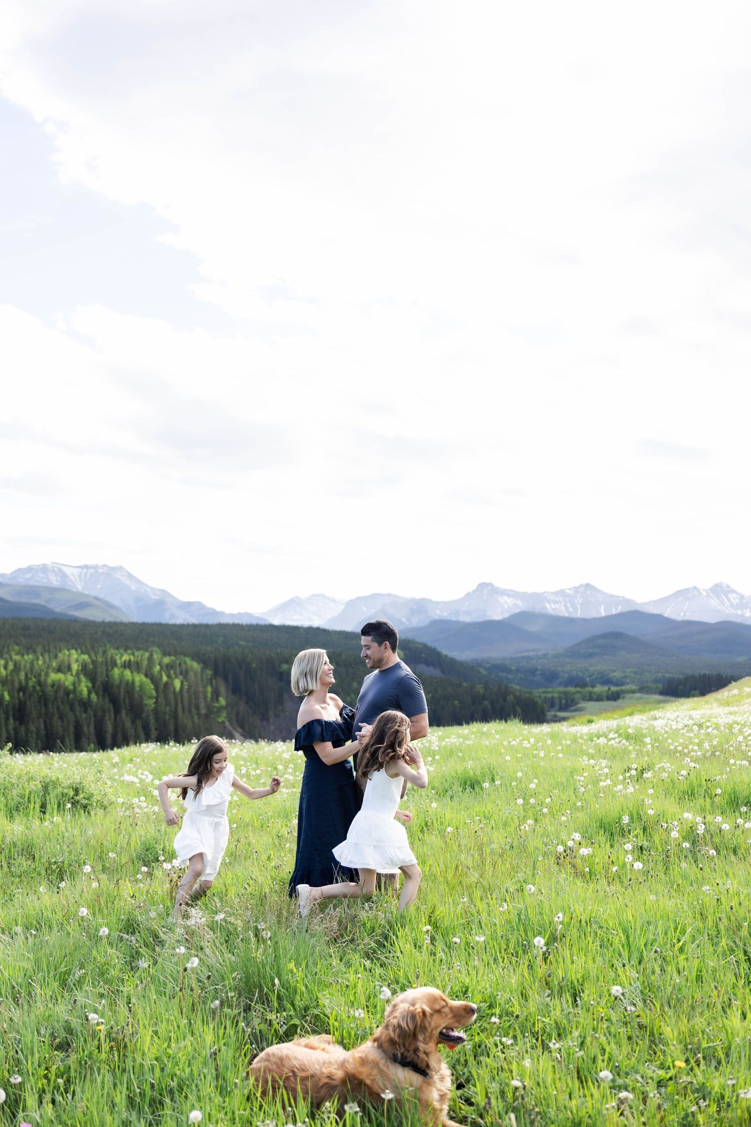 Family with two girls in white dresses and golden retriever enjoying a sunny mountain meadow with snow-capped peaks.