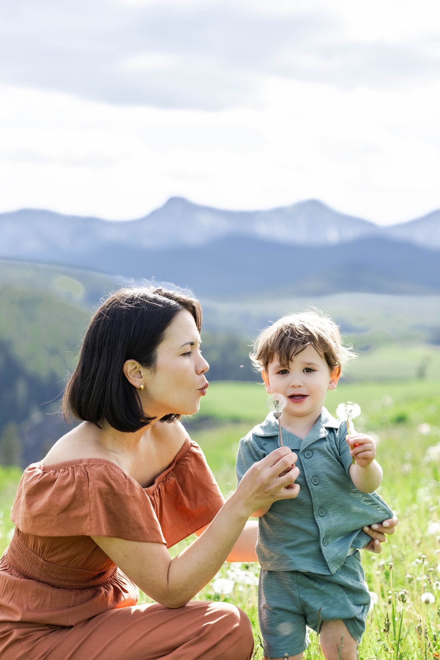 Mother and toddler blowing dandelion seeds in a green meadow with mountain backdrop on a cloudy day.