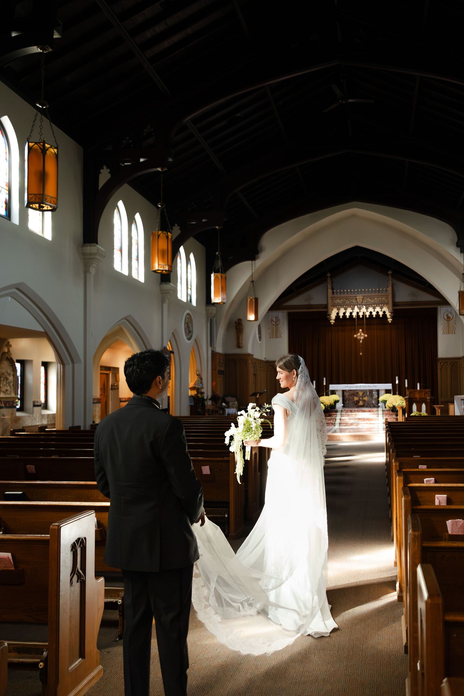 Bride and groom standing in elegant church aisle, bride in white gown with veil holding bouquet before ceremony.