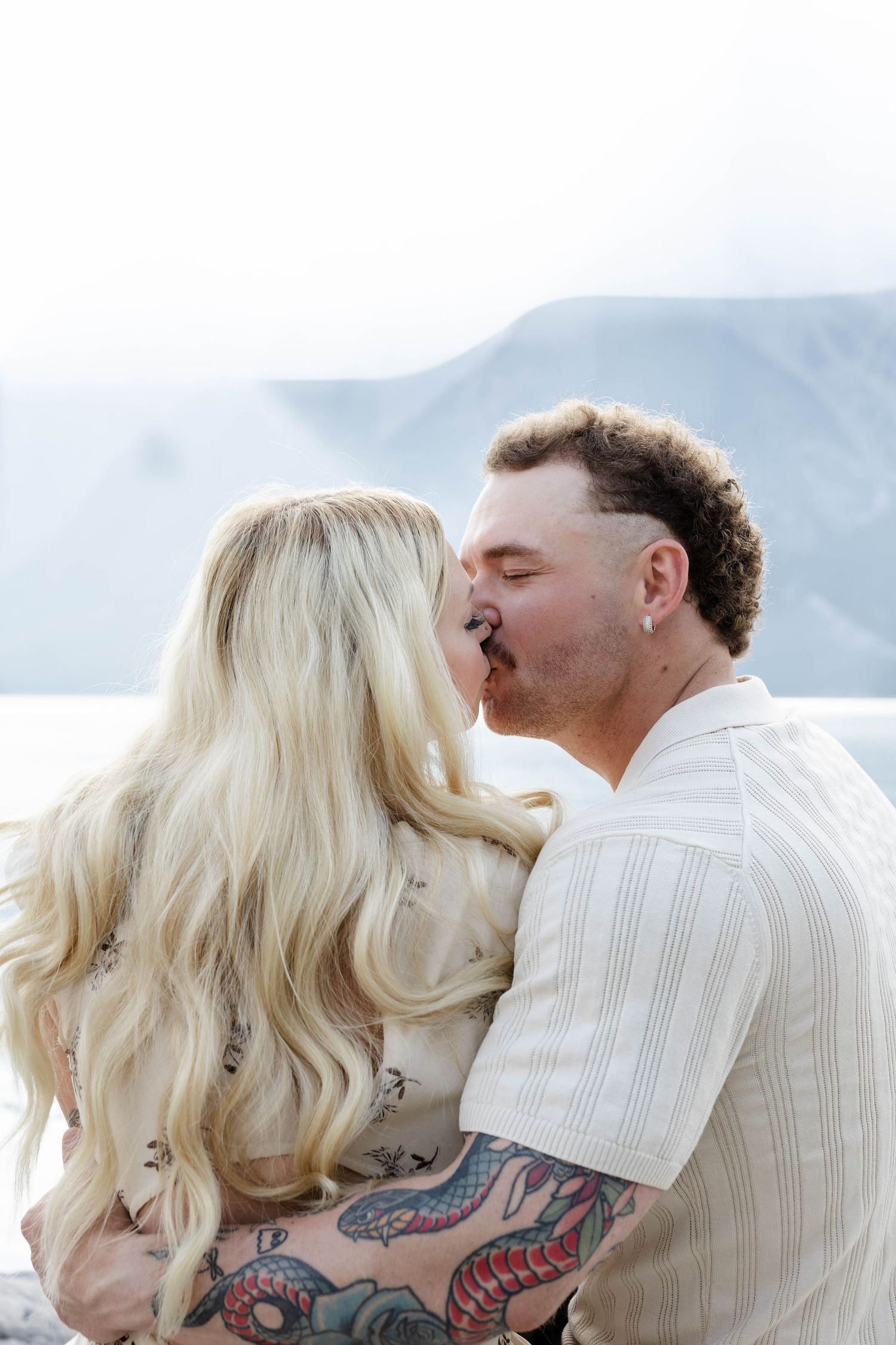 Couple sharing a romantic kiss outdoors with mountain backdrop, man with tattoos embracing blonde woman.Romantic couple in Minnewanka Lake i
