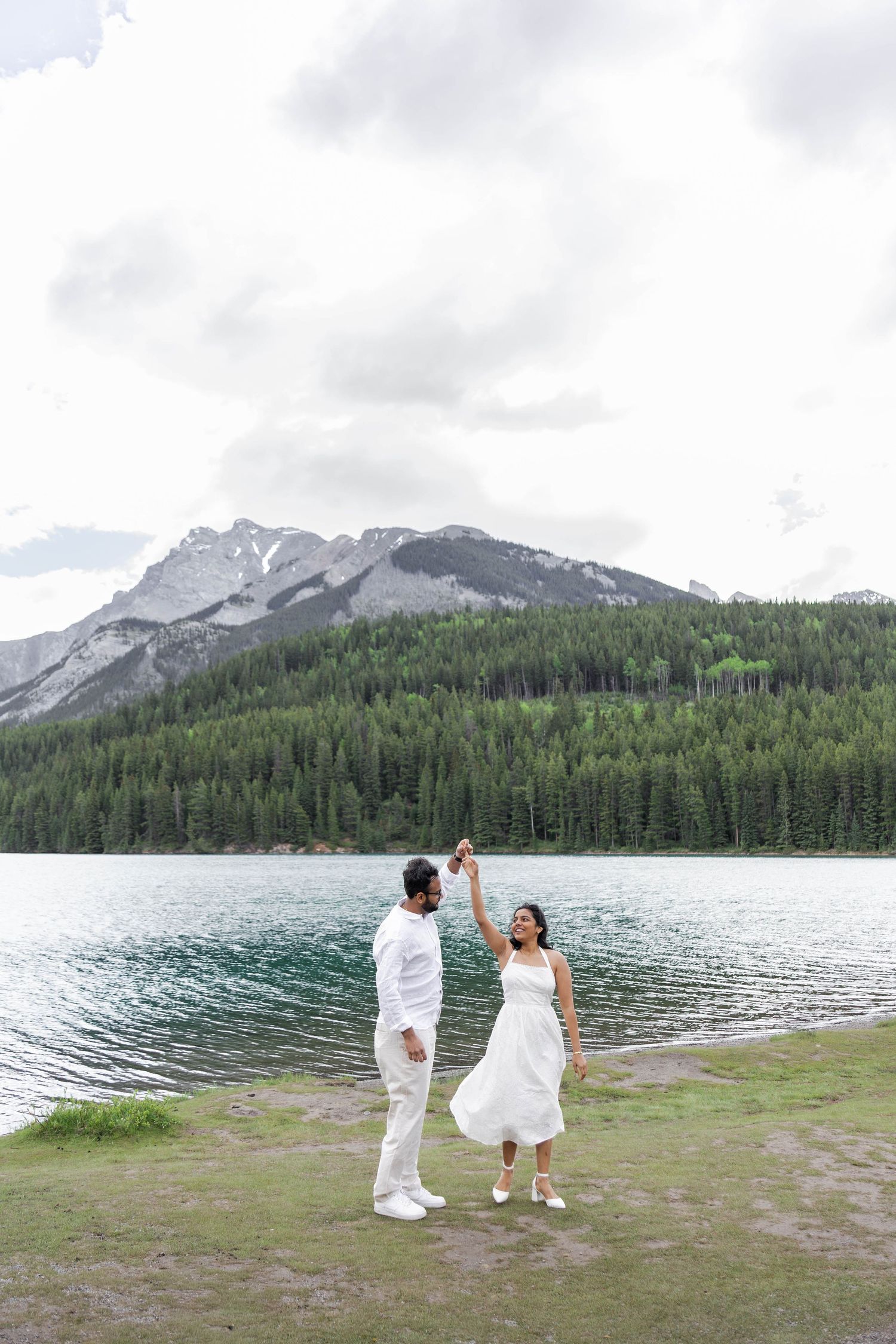 Couple dancing in white outfits by a turquoise lake with mountain and forest backdrop on a cloudy day.