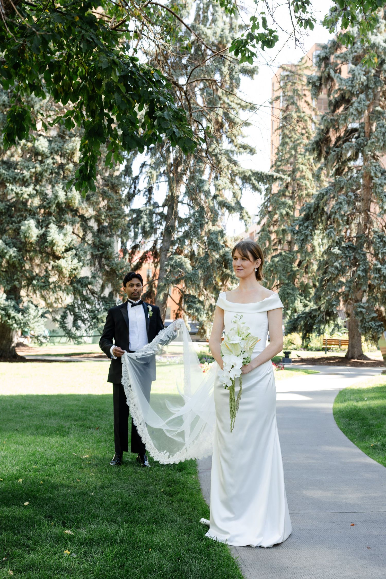 Bride walks park path in white gown holding bouquet while groom carries her veil behind her.