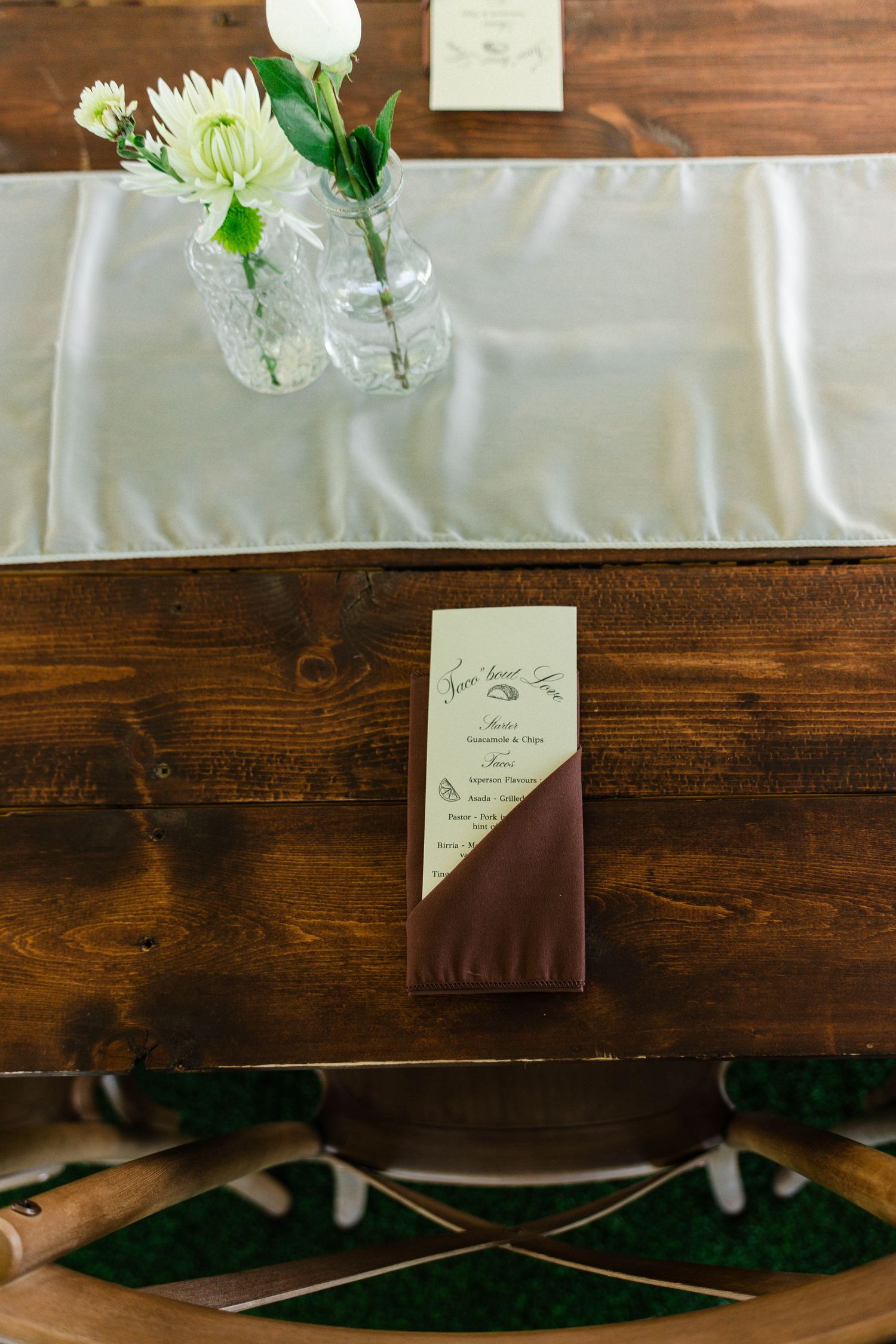 Rustic wooden wedding table with gray satin runner, white floral centerpiece in glass vases, and elegant burgundy menu card.