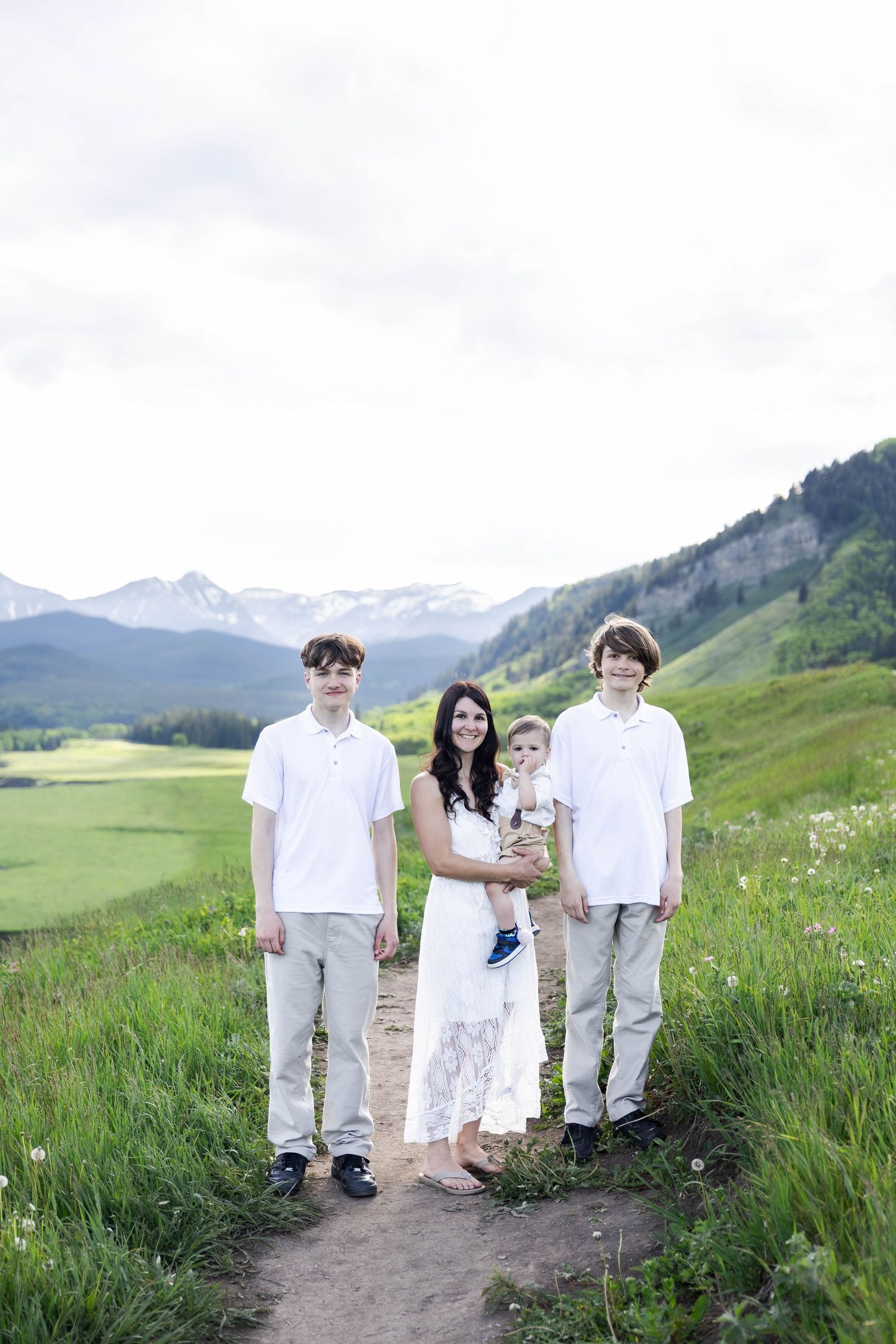 Family of four posing on a mountain trail surrounded by lush green meadows and snow-capped peaks.