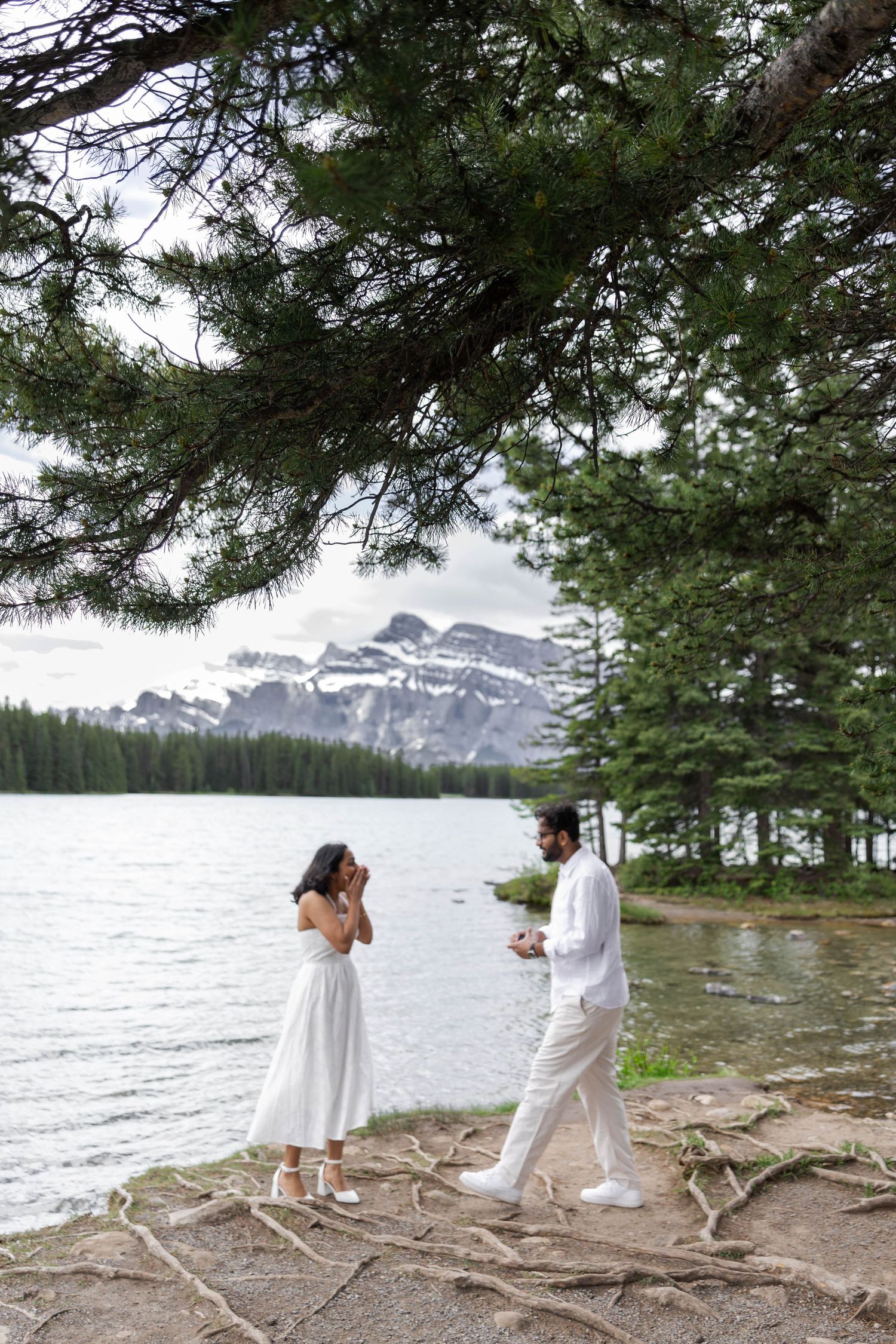 Couple in white outfits standing by a mountain lake with snow-capped peaks and pine trees in Banff.
