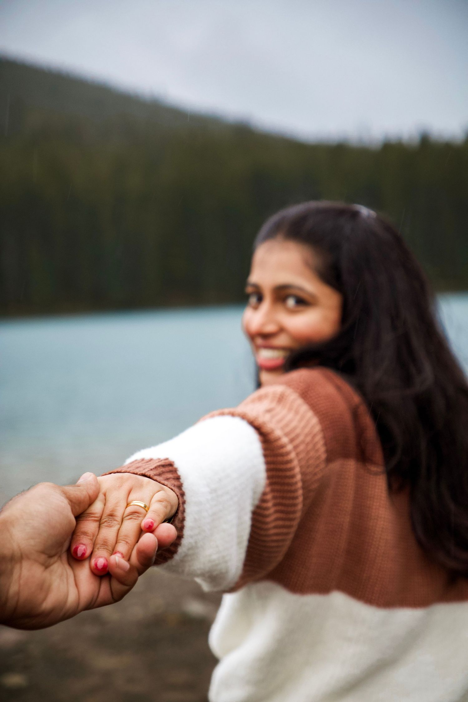 Smiling woman in brown sweater holds hands with partner near a serene blue lake surrounded by forest.