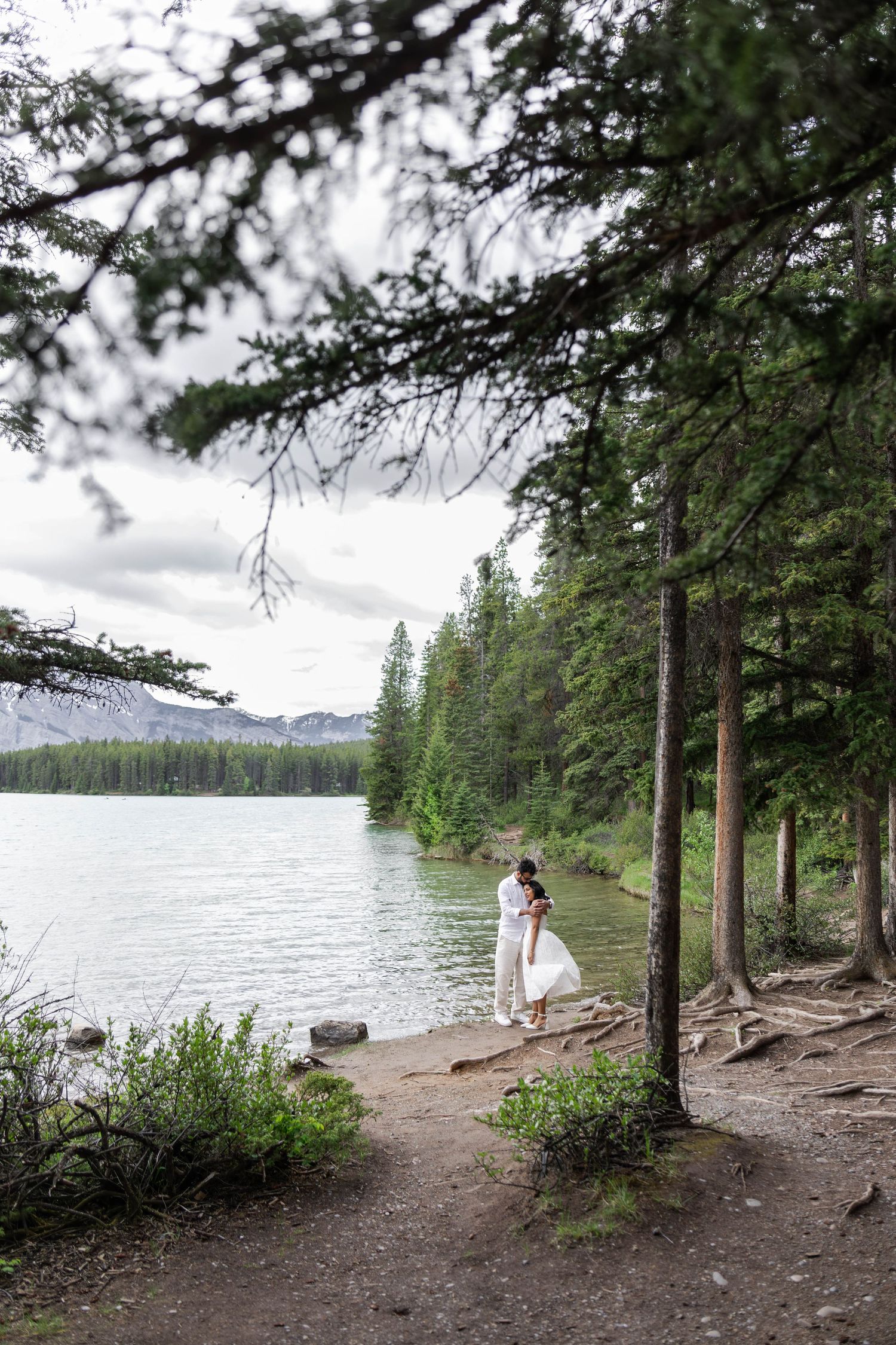 Couple embracing near a serene mountain lake in Banff, framed by tall pine trees and lush greenery.