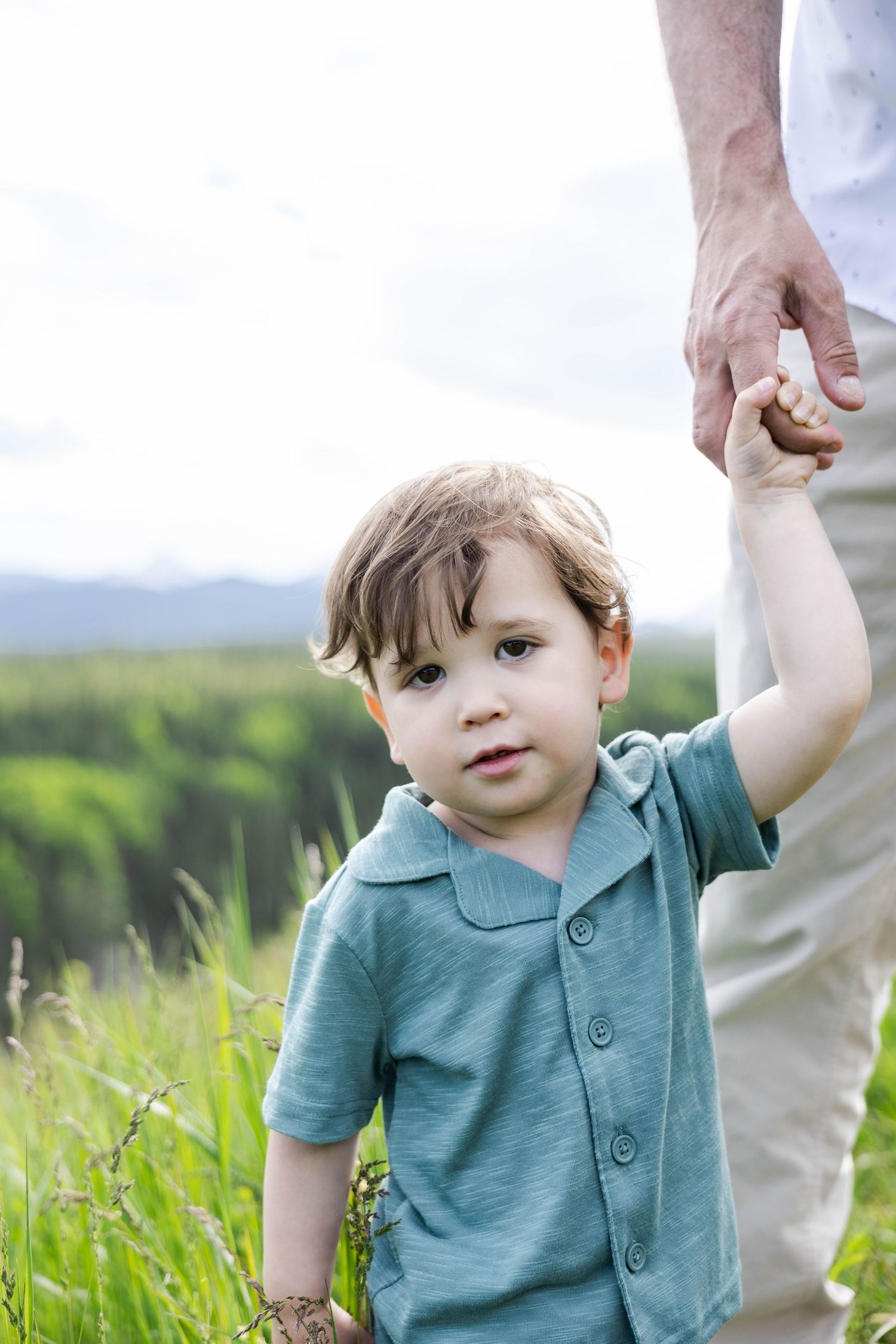 A toddler boy in a teal shirt holds an adult's hand while standing in a lush green meadow outdoors.
