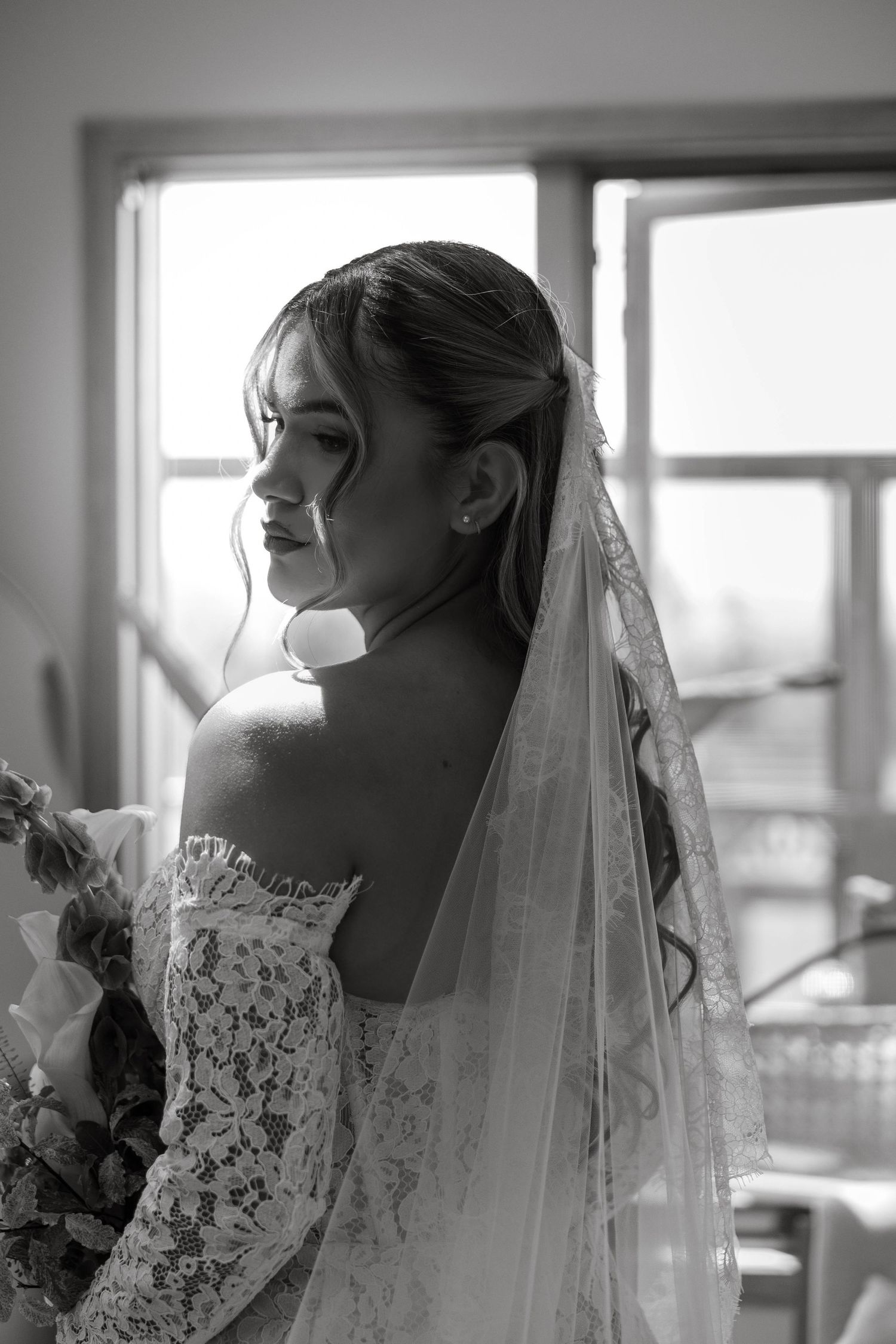 Black and white portrait of a bride in lace off-shoulder gown and veil, gazing down near a bright window.