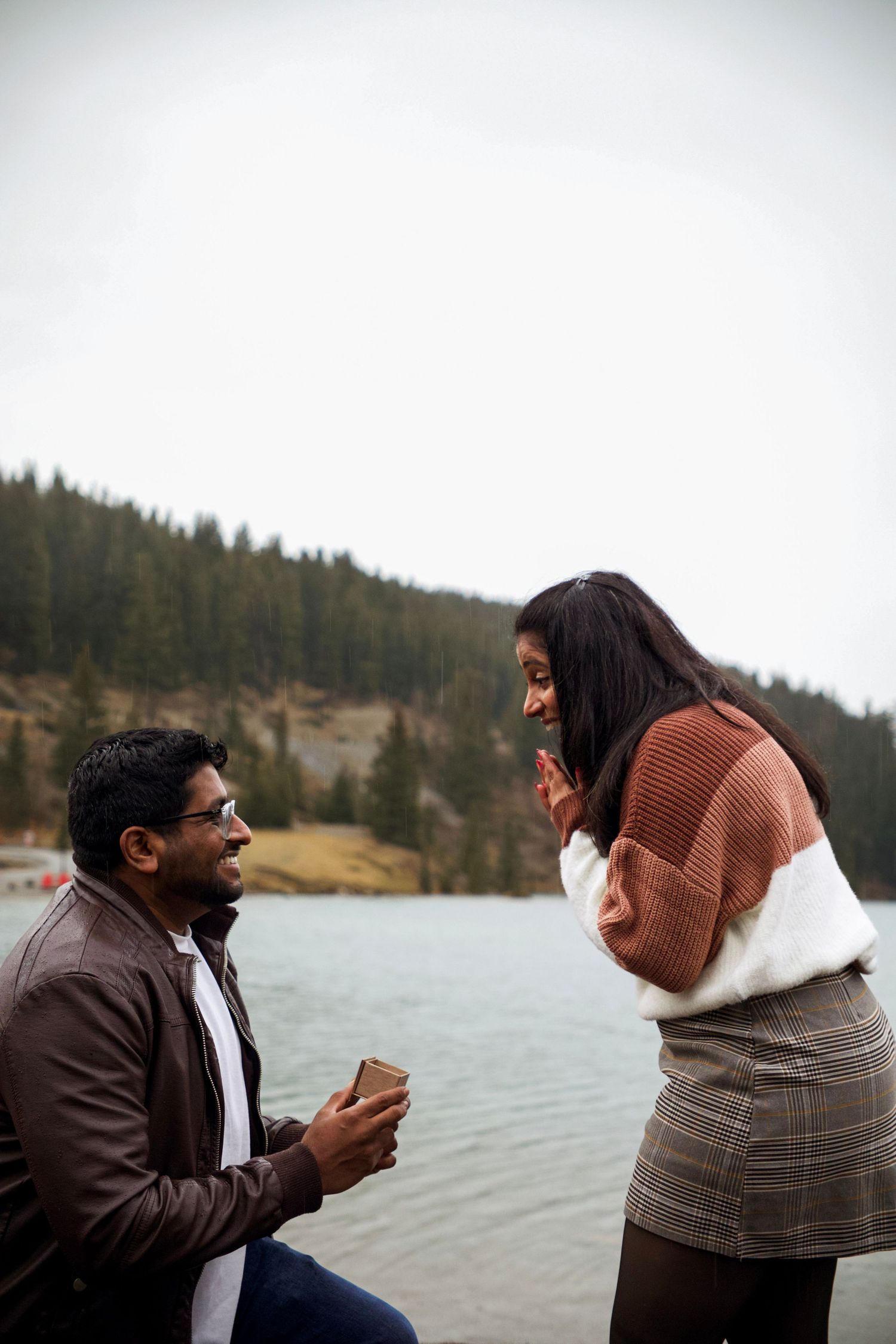 Man proposes to woman by a mountain lake, kneeling with ring box as she covers her mouth in surprise in Banff
