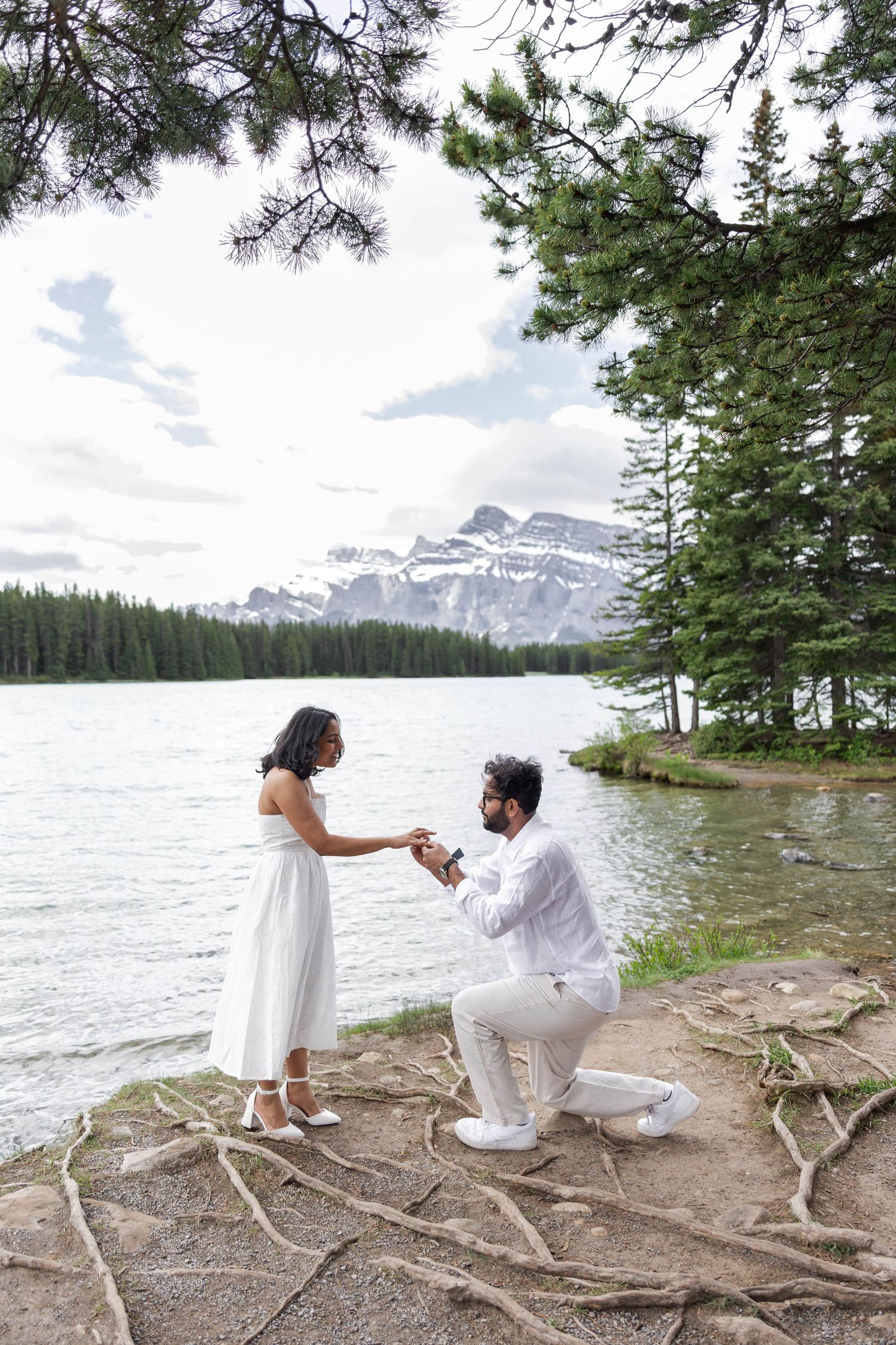 Man proposing to woman by a mountain lake surrounded by pine trees and snow-capped peaks in Banff, Canada.