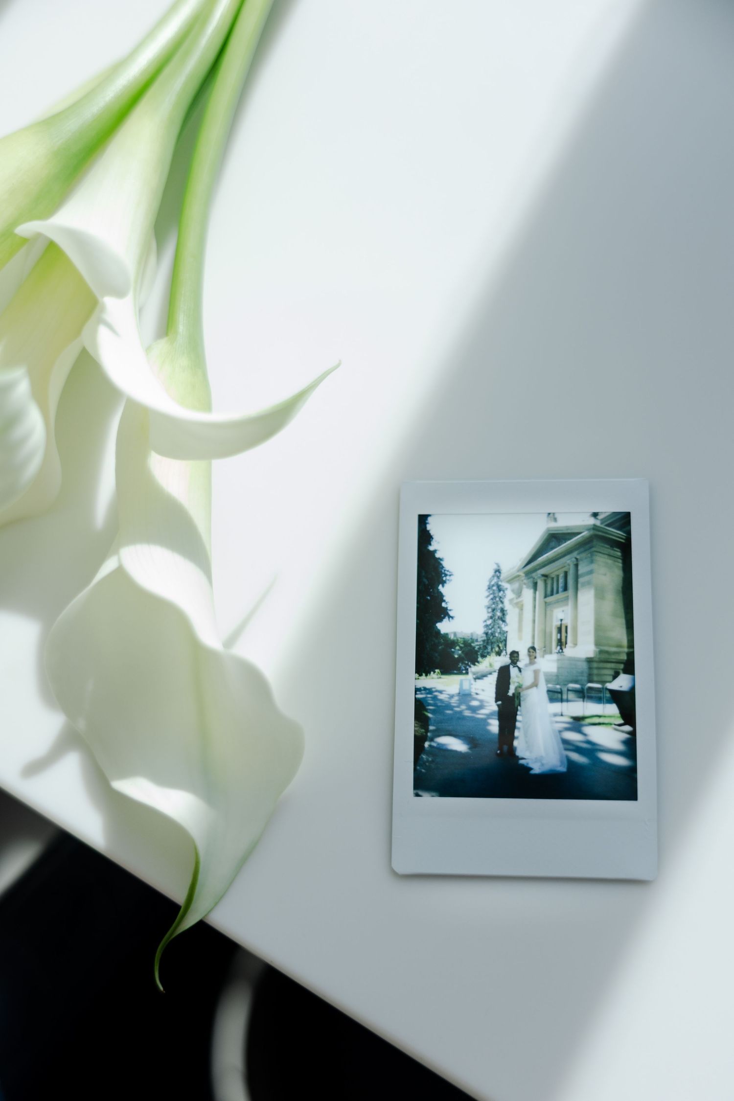A Polaroid wedding photo beside white calla lilies on a white surface in natural light.