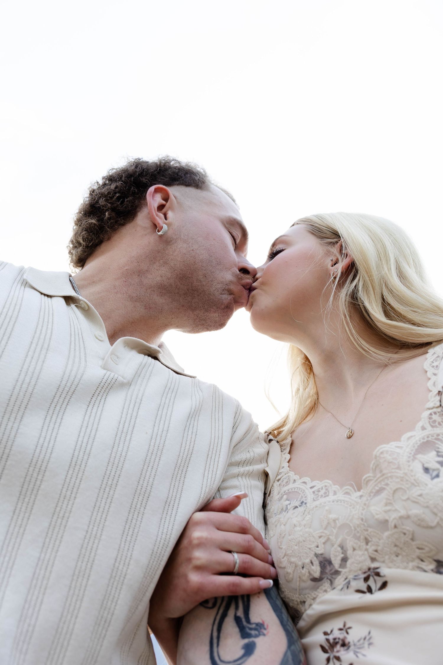 A couple shares a romantic kiss, the groom in a striped suit and bride in lace dress with tattoos visible.Romantic couple in Minnewanka Lake