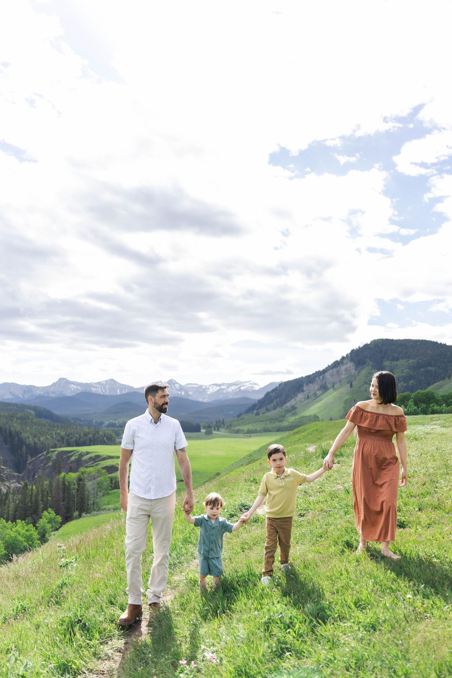 Family of four holding hands walking through a lush green mountain meadow on a cloudy summer day.
