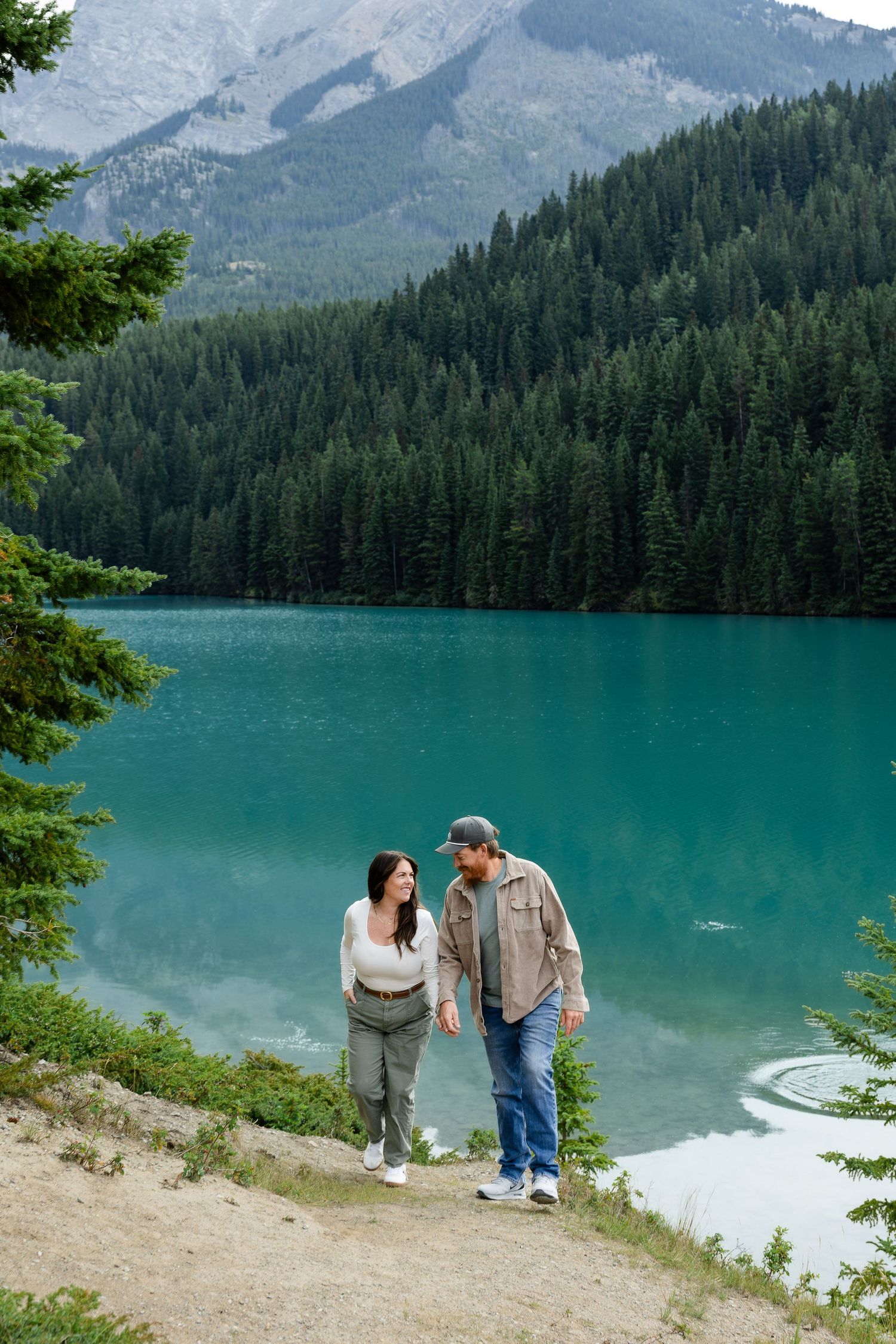 Couple walking hand in hand by a turquoise alpine lake surrounded by pine forests and mountains.