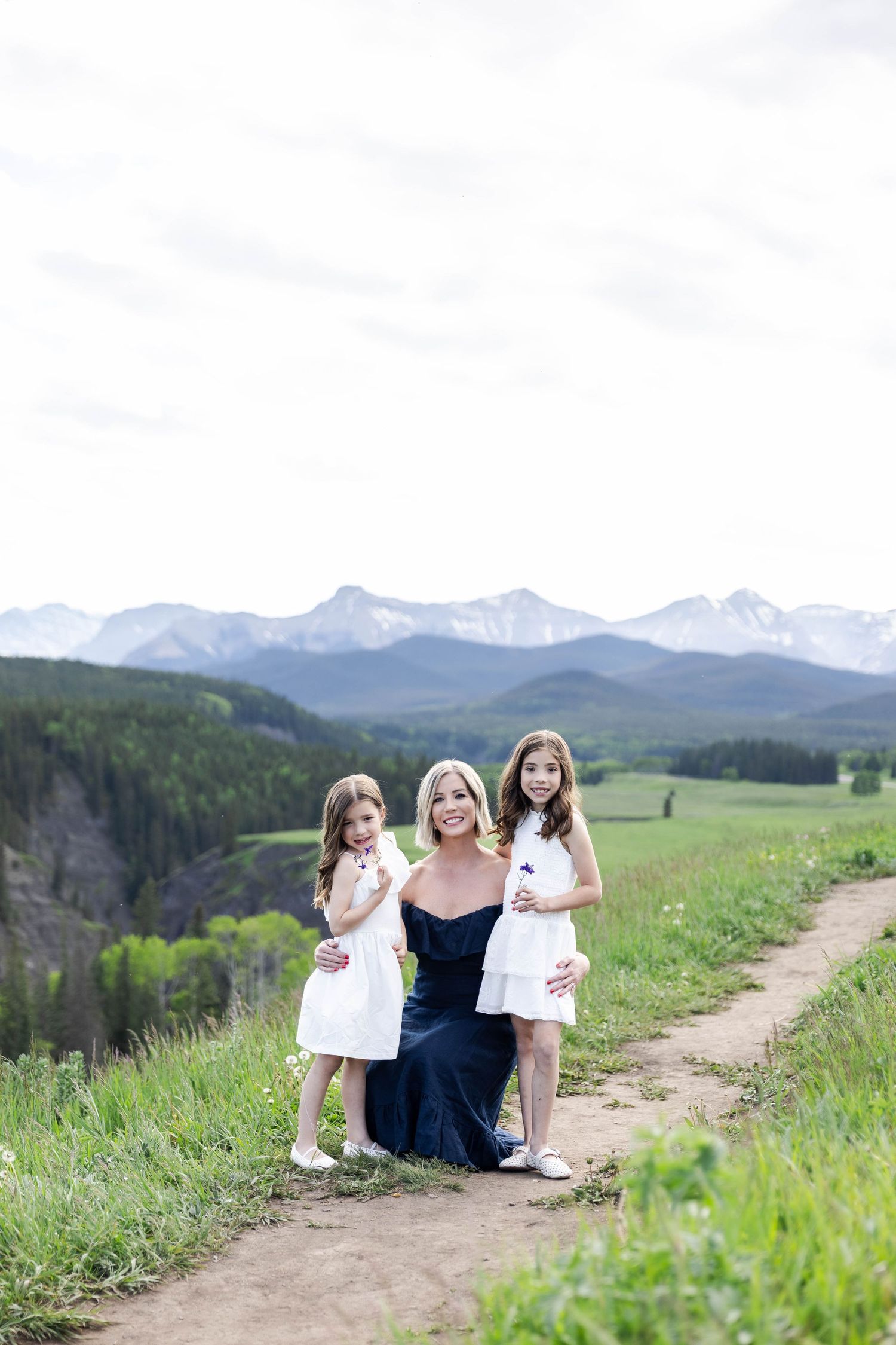 A woman in a navy dress kneels with two girls in white dresses on a mountain trail with snowy peaks behind them.