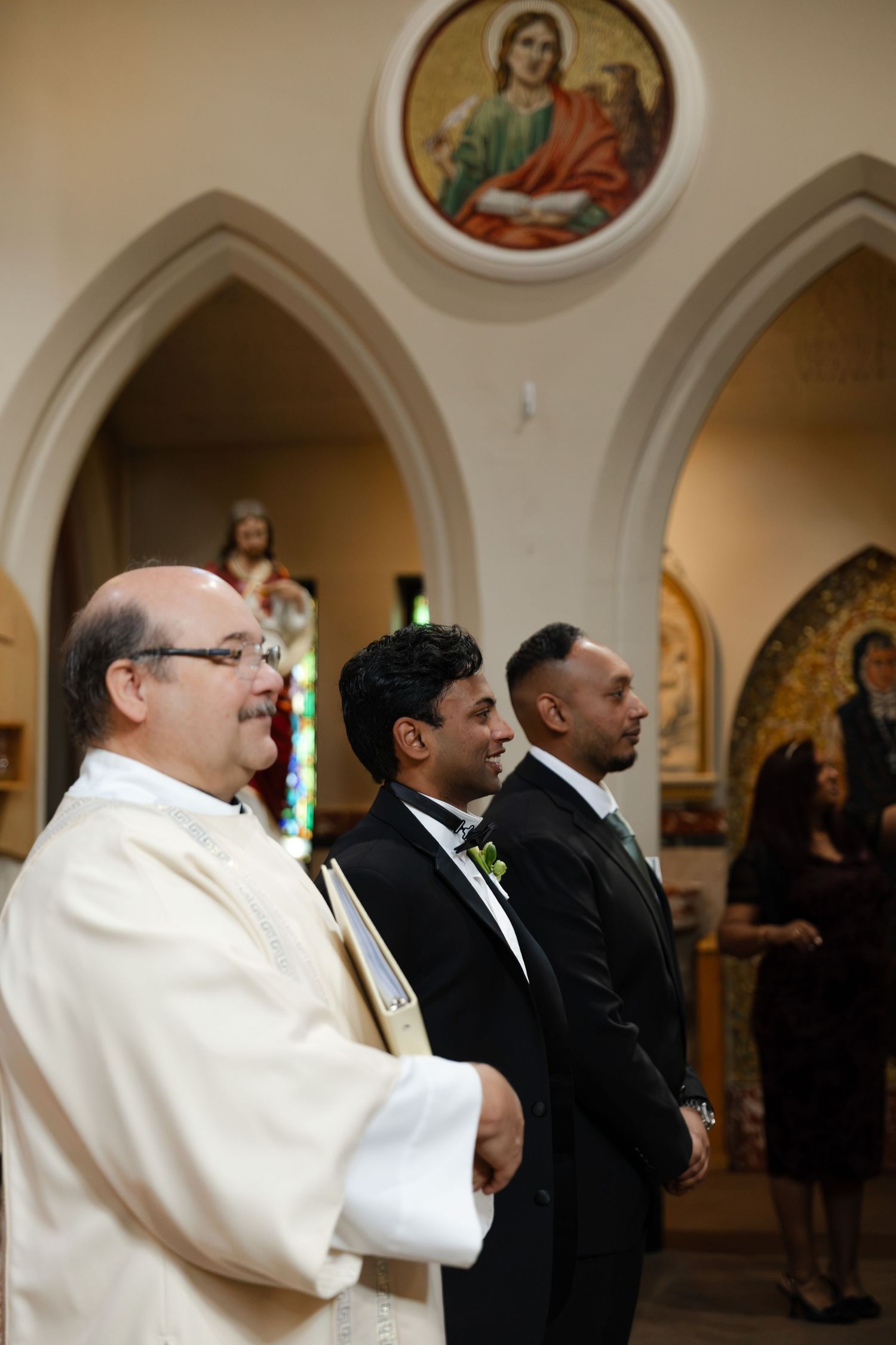 Priest and groomsmen standing at church altar during wedding ceremony with religious mosaic artwork in background.