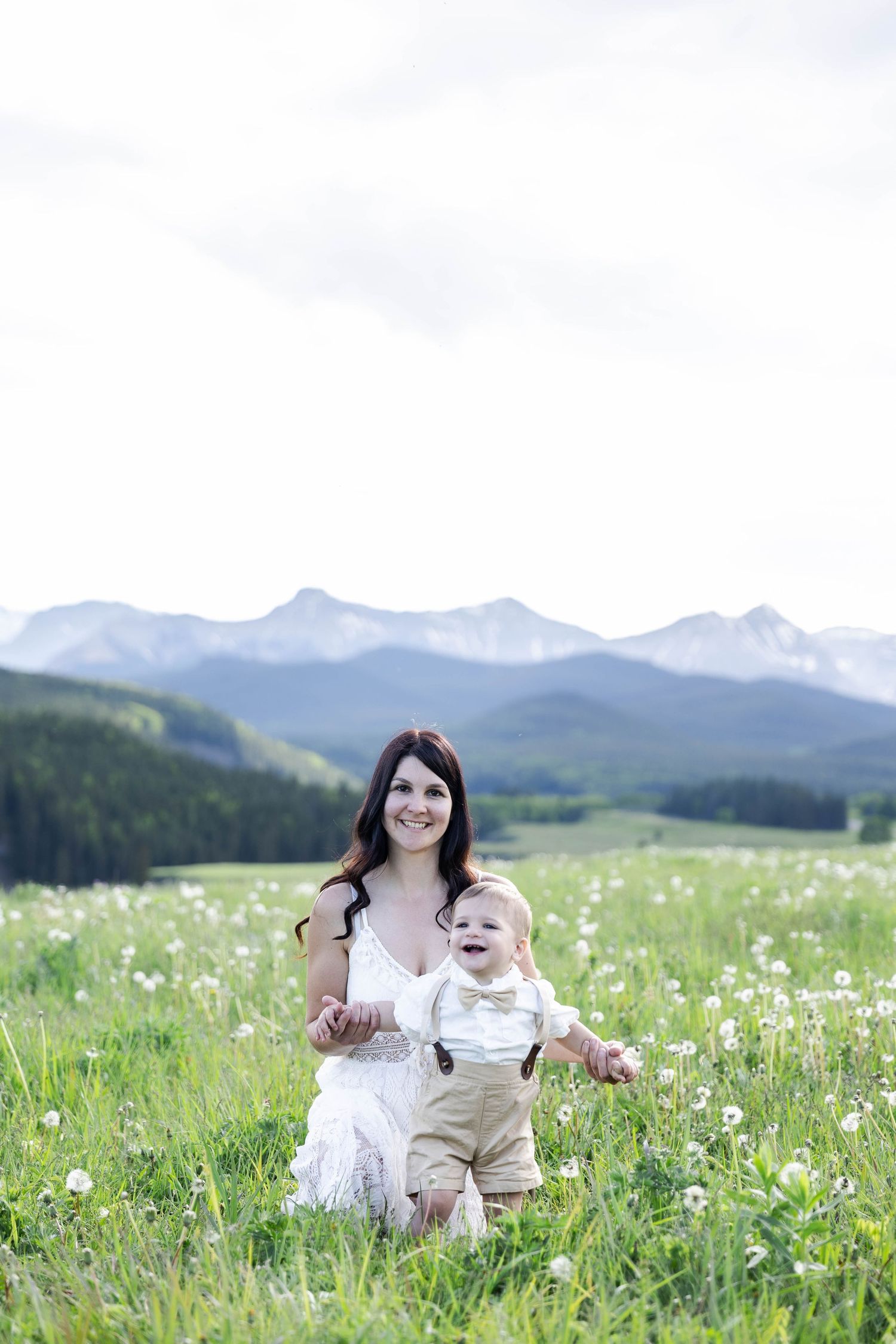 A smiling mother in a white dress helps her laughing baby stand in a green mountain meadow with snow-capped peaks behind them.