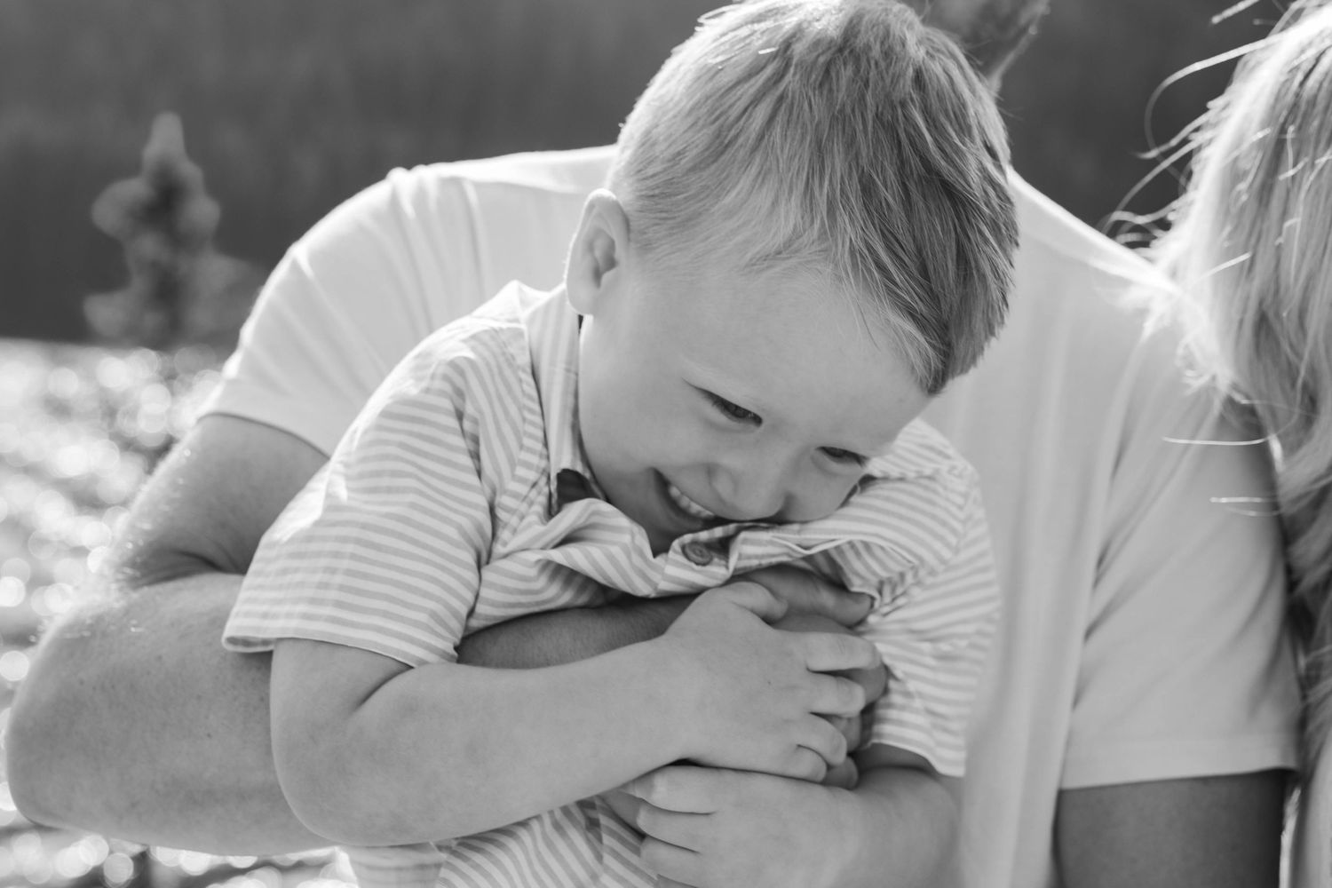 Black and white photo of a smiling toddler boy being held and cuddled by his father outdoors.in Banff