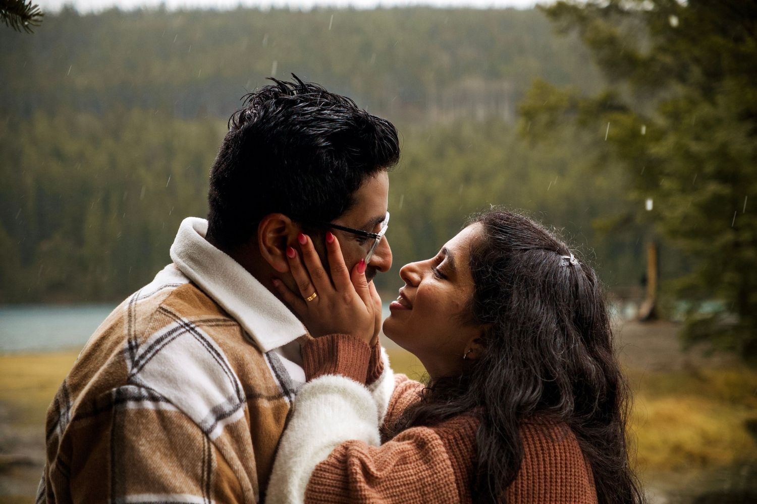 Couple sharing a romantic moment outdoors near a lake surrounded by pine trees in a cozy autumn setting.