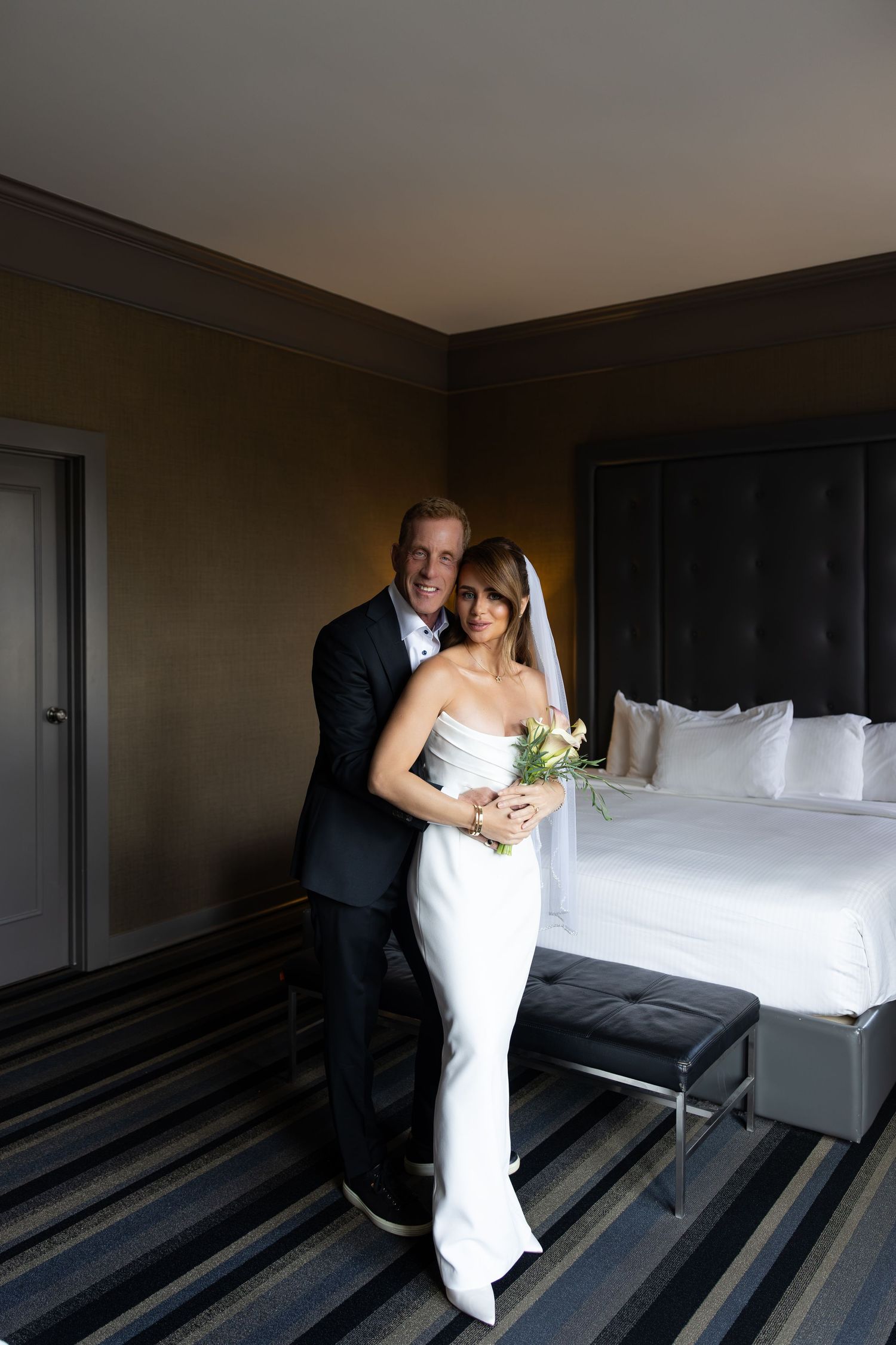 Bride in white strapless gown with veil holding bouquet embraced by groom in dark suit in elegant hotel room.