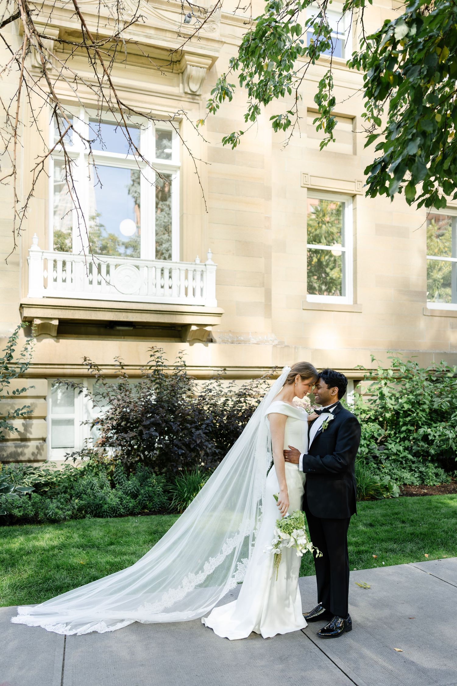 Bride and groom share an intimate moment outside an elegant stone mansion, her cathedral veil trailing on the lawn.
