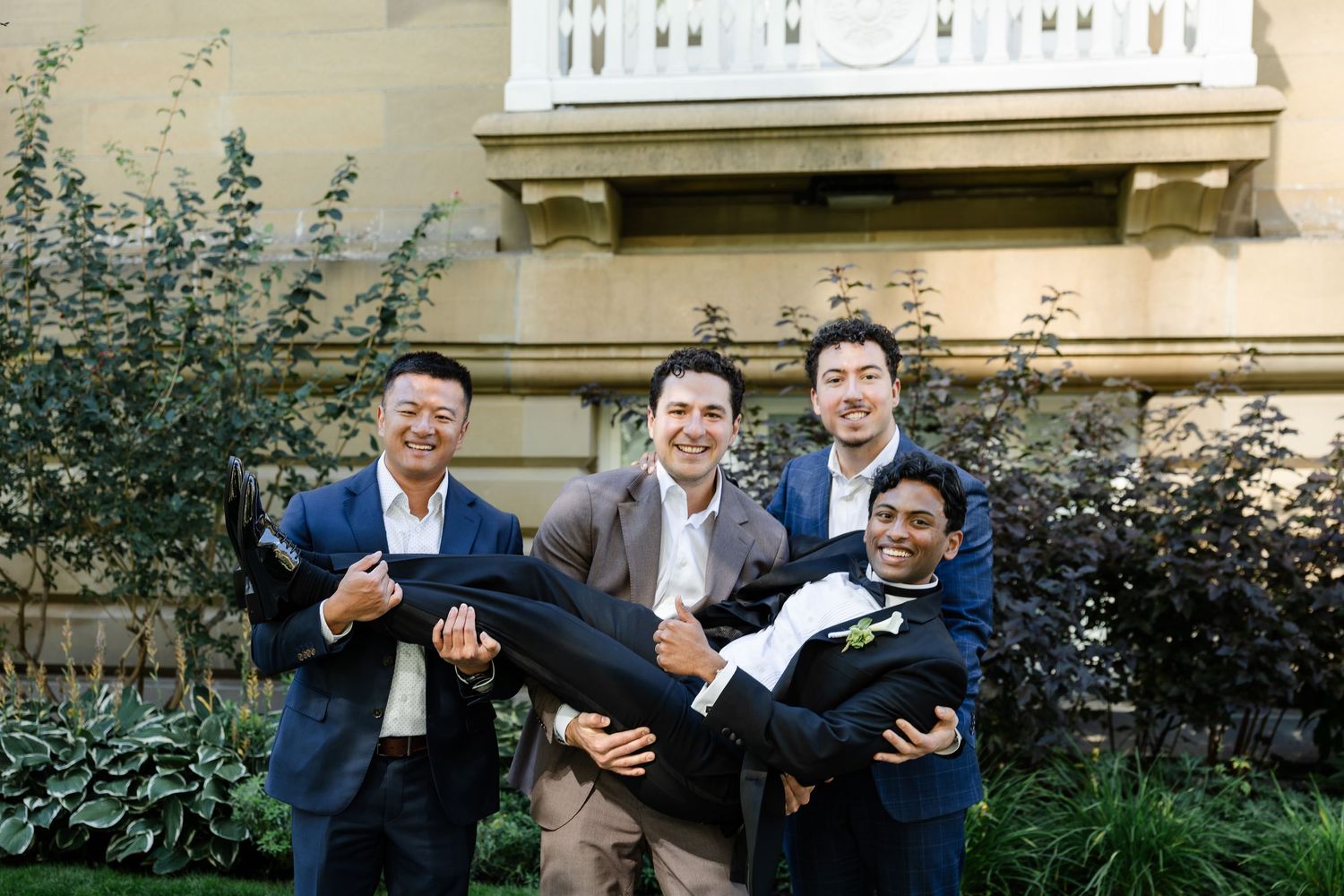 Three groomsmen joyfully lift the groom in front of an elegant stone building surrounded by lush greenery.