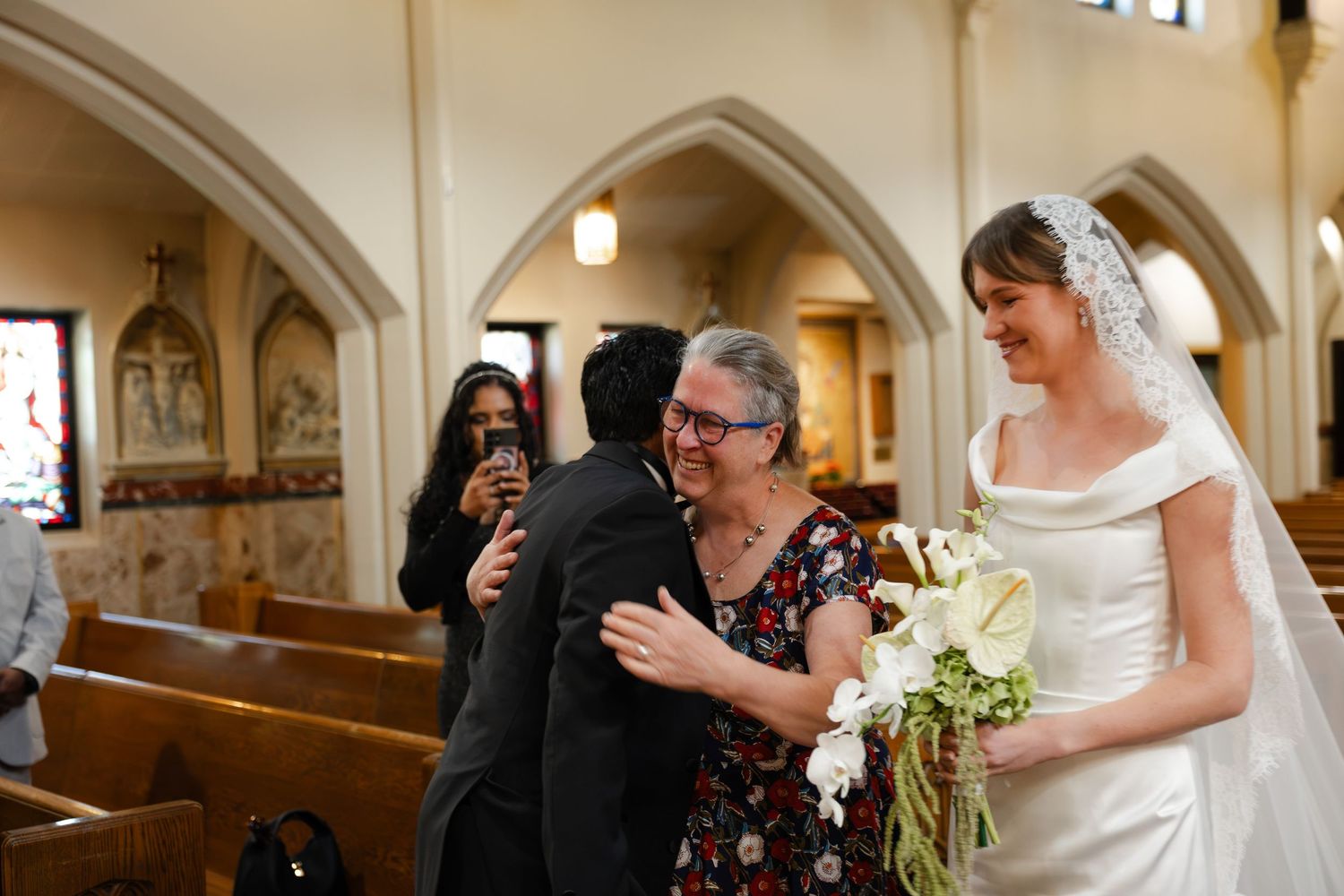 Bride in white gown holding bouquet hugs guest inside church during wedding ceremony celebration.