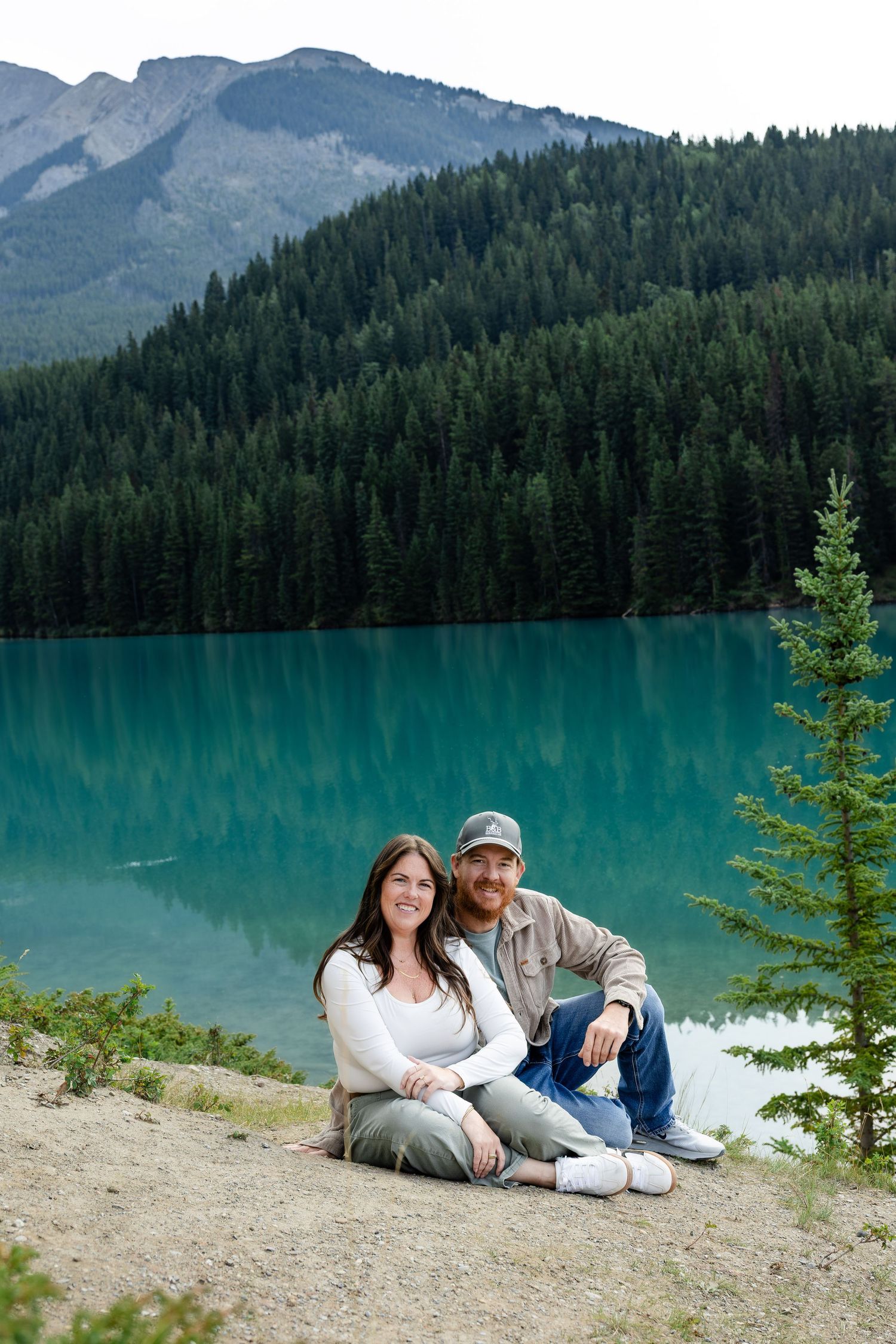 Couple sitting together on rocky shore of turquoise mountain lake surrounded by evergreen forest and peaks.