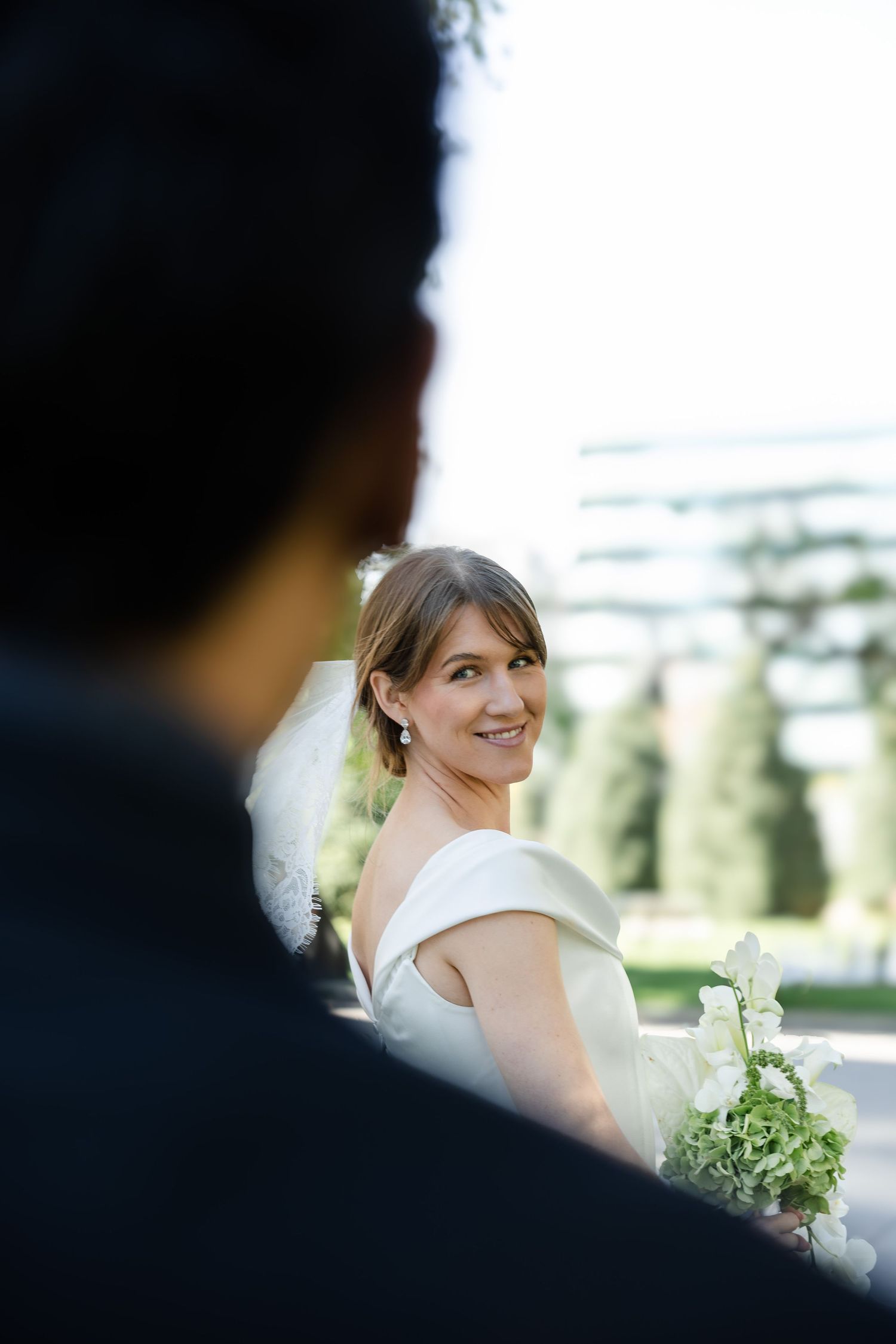 Bride in white off-shoulder gown smiles back at groom, holding bouquet, outdoors at wedding ceremony.