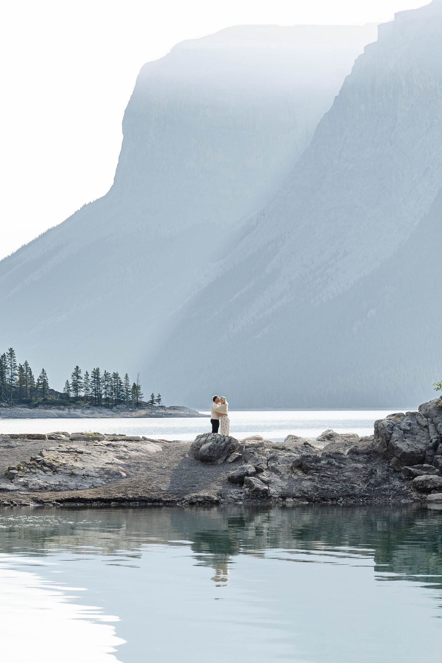Couple embracing on rocky shore of a misty mountain lake with calm reflective water and evergreen trees.Romantic couple in Minnewanka Lake i