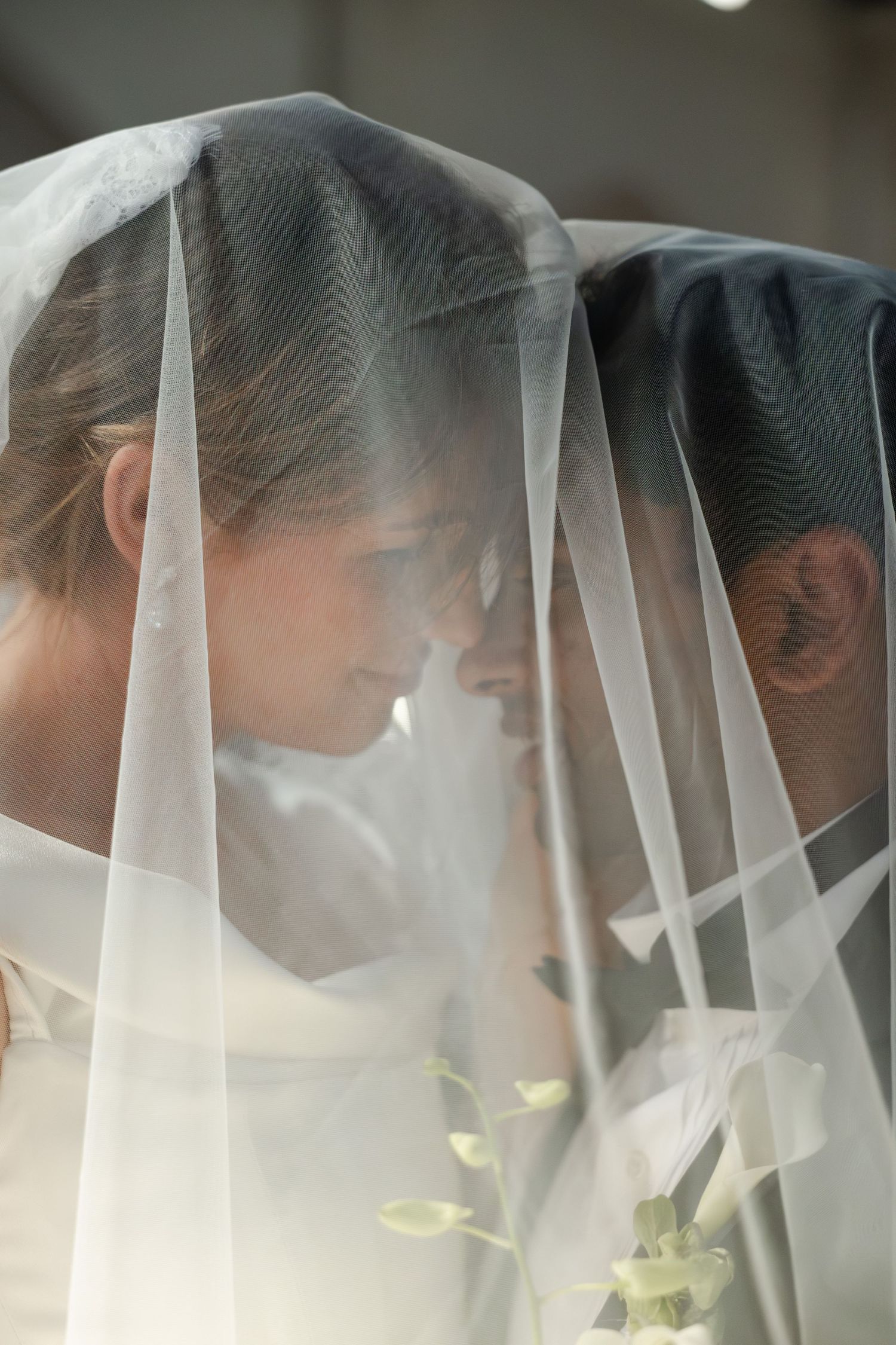 Bride and groom share an intimate moment under a flowing white wedding veil, foreheads touching tenderly.