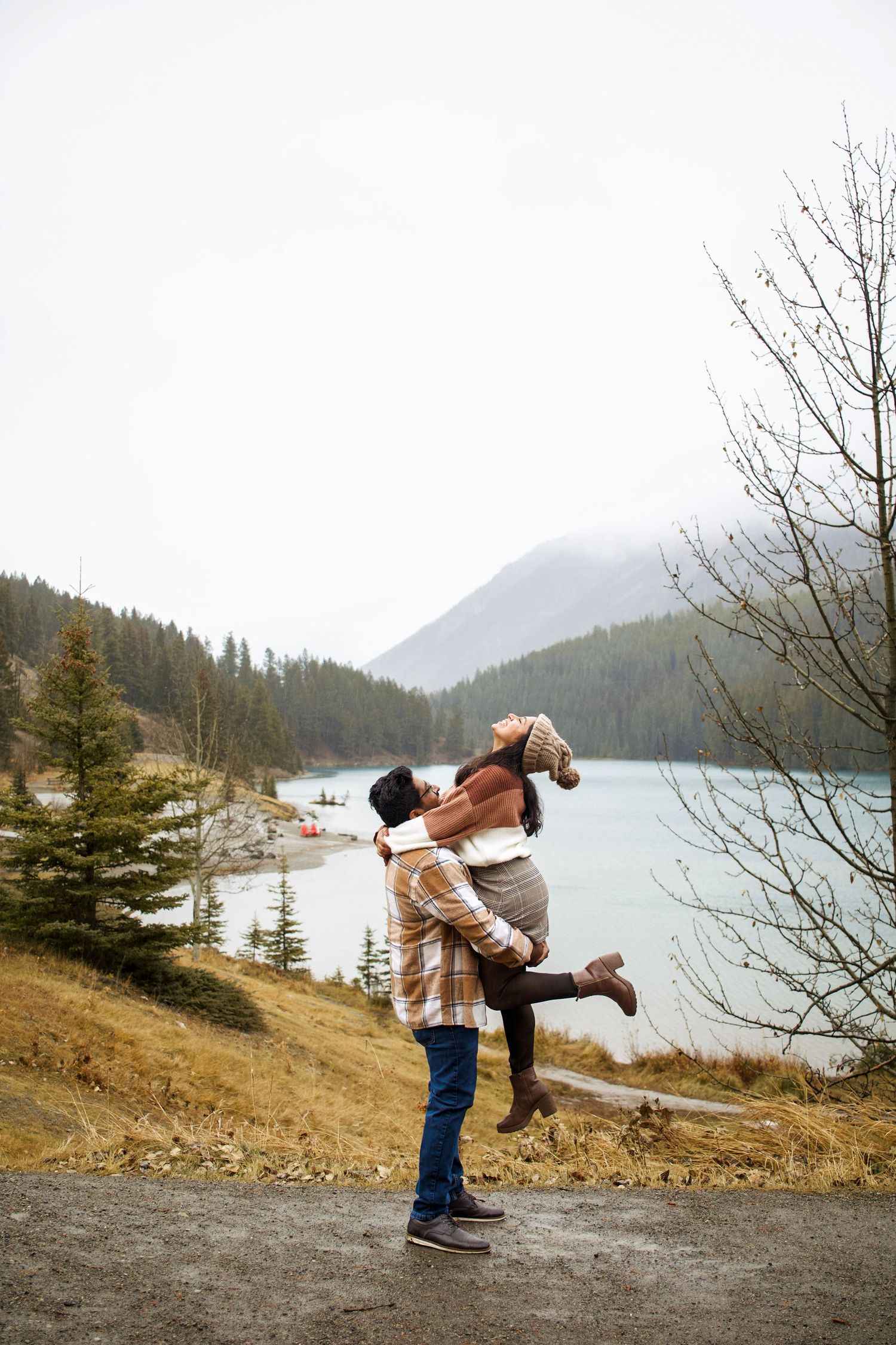 Couple embracing by a mountain lake, man lifting woman joyfully, surrounded by pine trees and misty peaks.