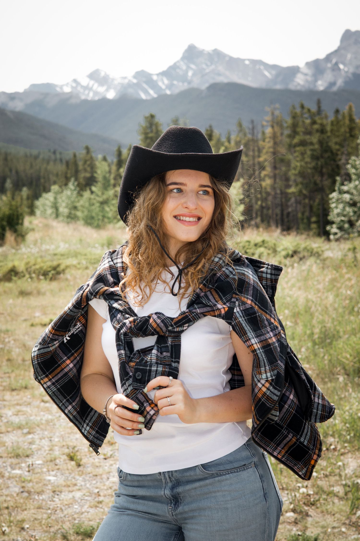 Smiling young woman in cowboy hat and plaid shirt holds camera in mountain meadow with pine trees and snowy peaks.