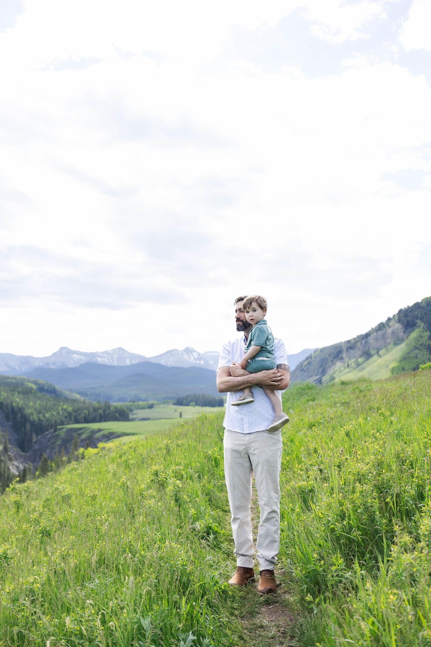 Father holding young child on a scenic mountain meadow trail with snow-capped peaks and green valley views.