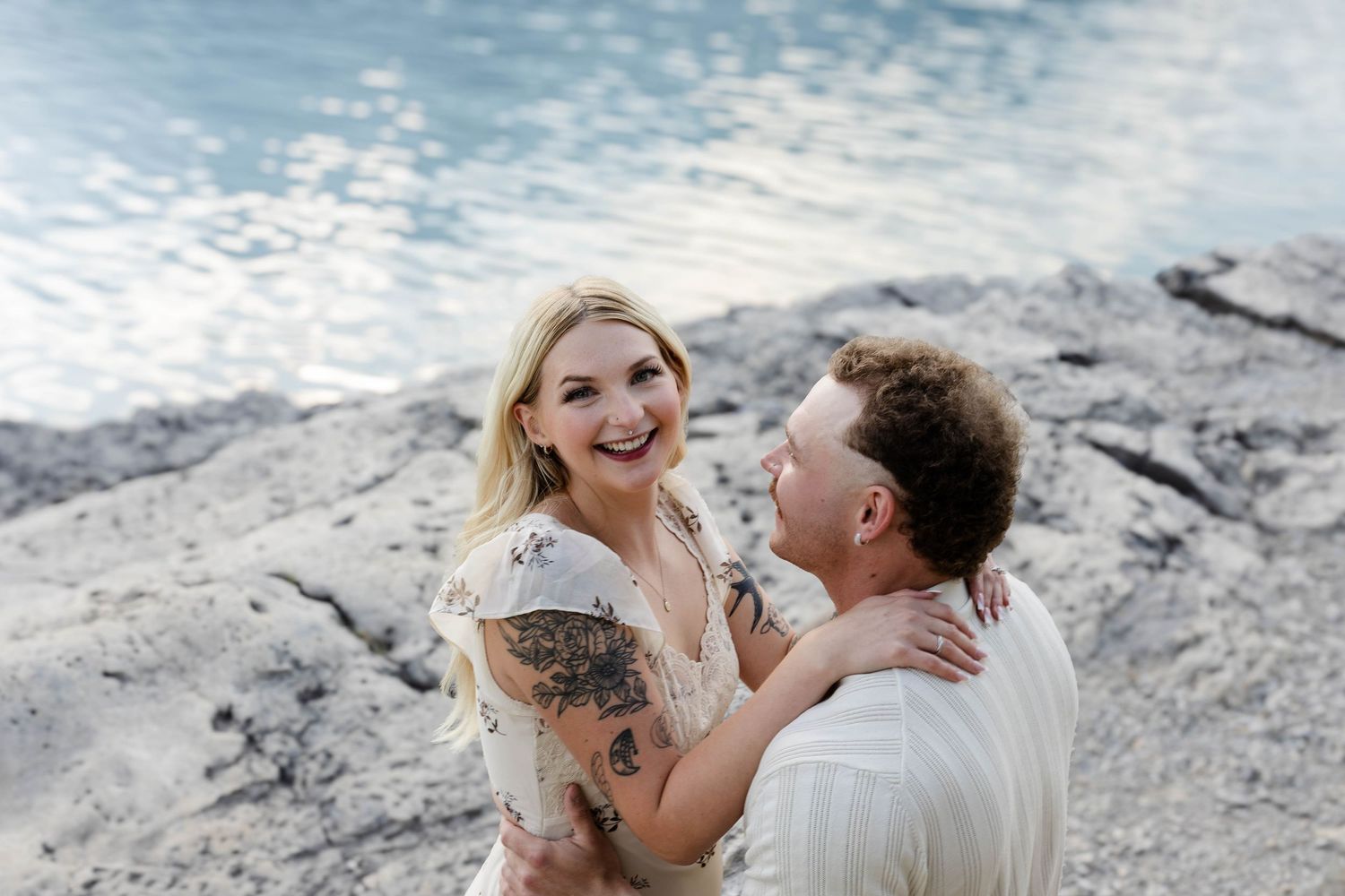 Couple laughing and embracing on sun-lit rocky lakeshore with sparkling blue water in background.Romantic couple in Minnewanka Lake in Banff