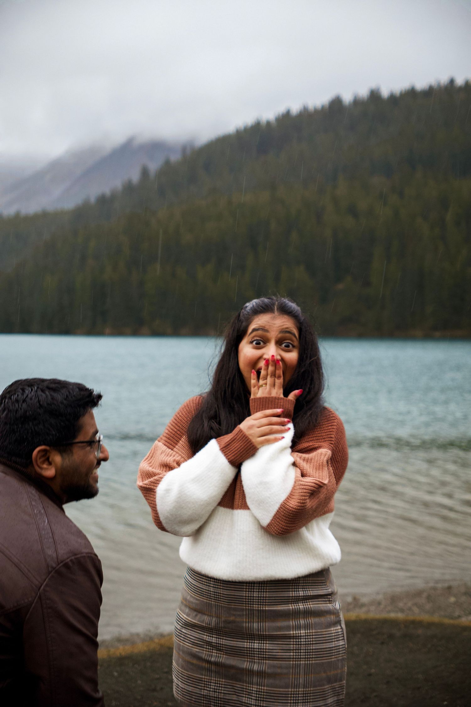 Woman reacts with surprise near a mountain lake as a man looks on, both standing outdoors in a scenic setting.