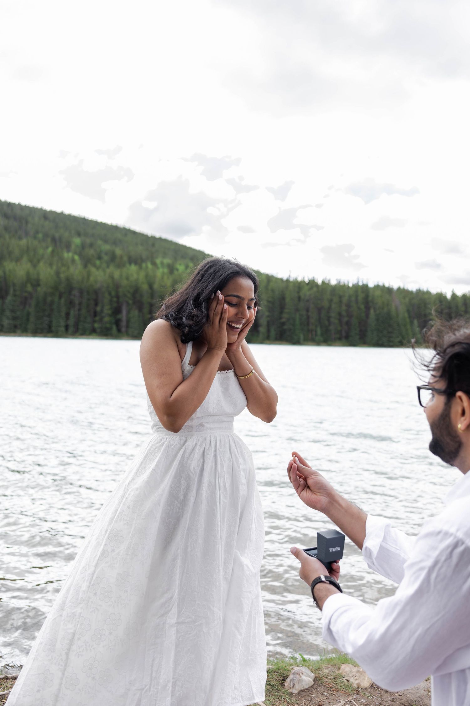 Man proposing to surprised woman in white dress by a scenic mountain lake surrounded by pine forest.
