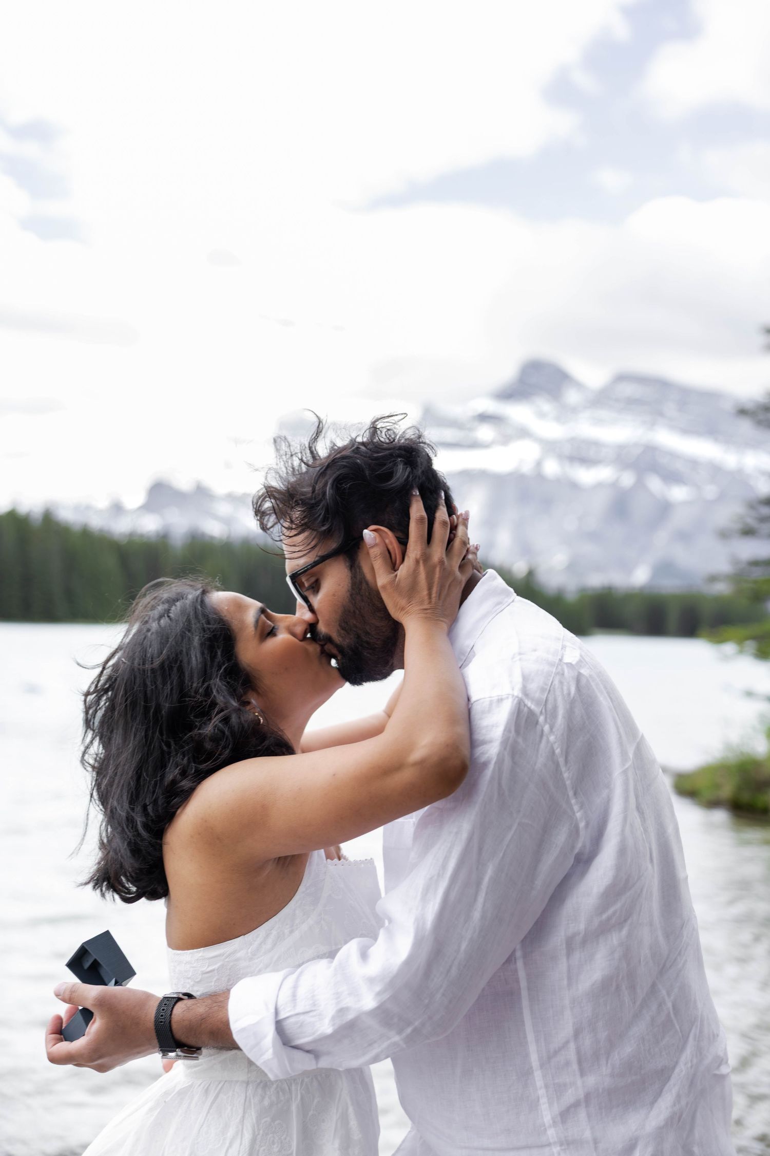 Romantic couple kissing by mountain lake, woman in white dress holding ring box during proposal.