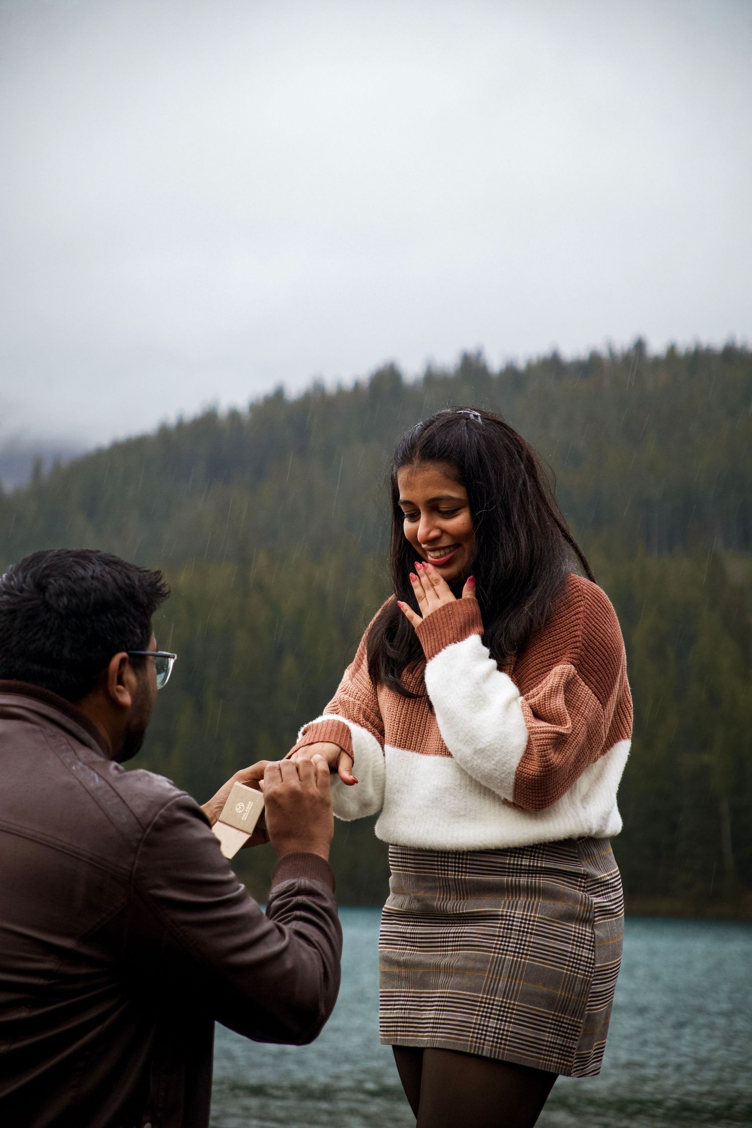 Man proposing to woman with ring box by a scenic mountain lake, her hand raised in surprise.