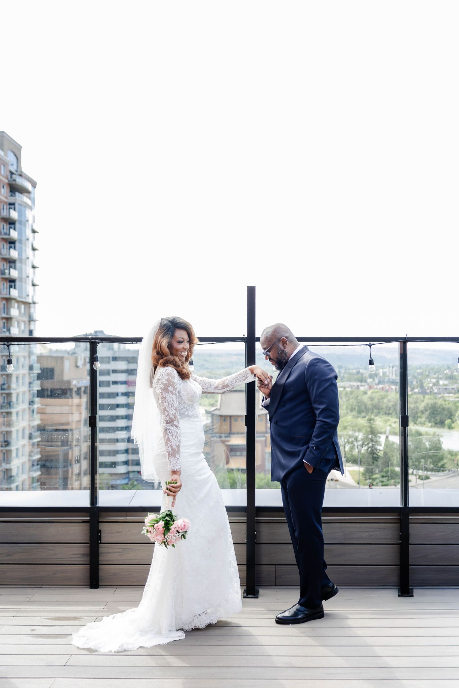 Groom kissing bride's hand on rooftop terrace with city skyline backdrop during outdoor wedding portrait.
