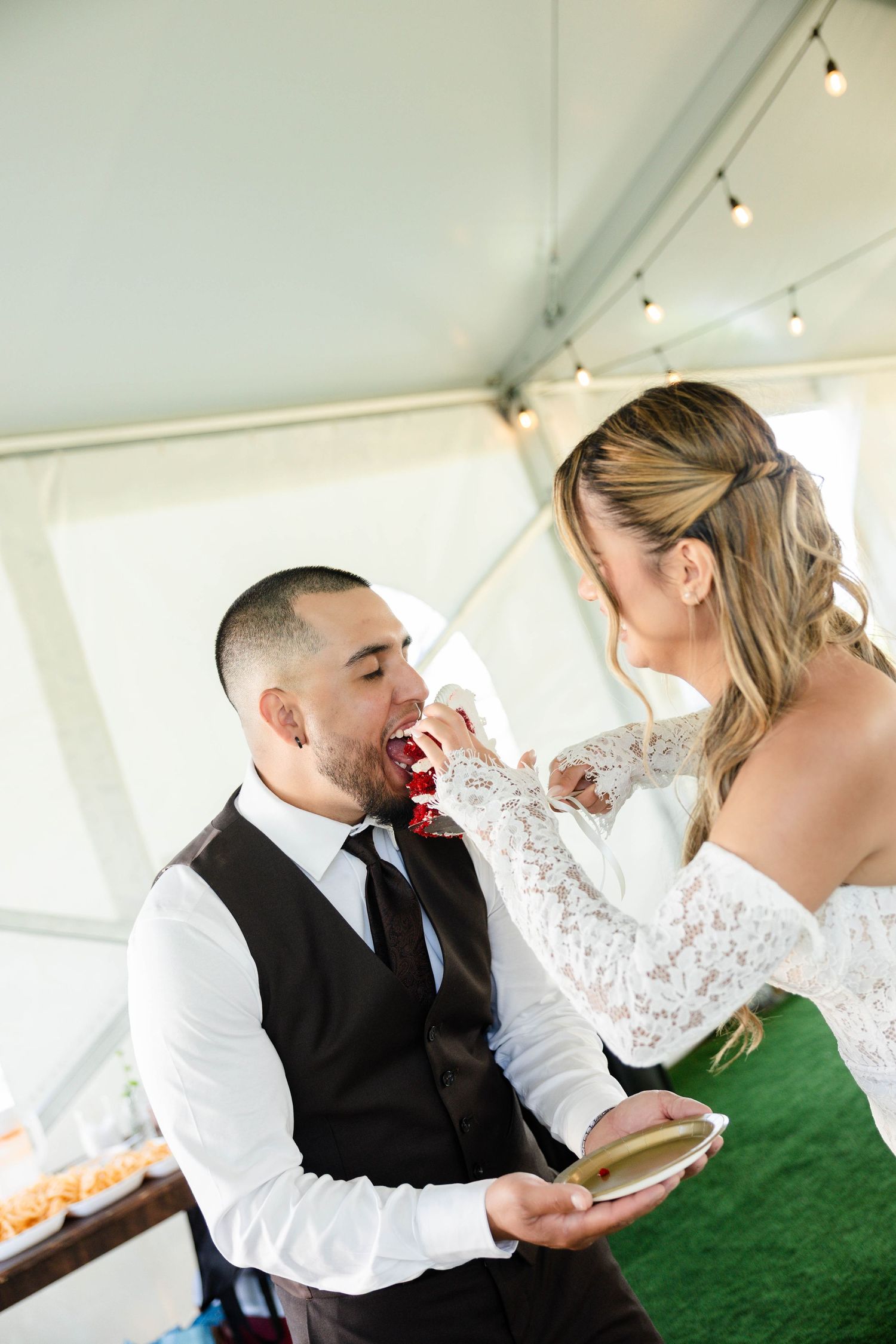 Bride in lace gown feeds wedding cake to groom in black vest and tie during outdoor tent reception celebration.