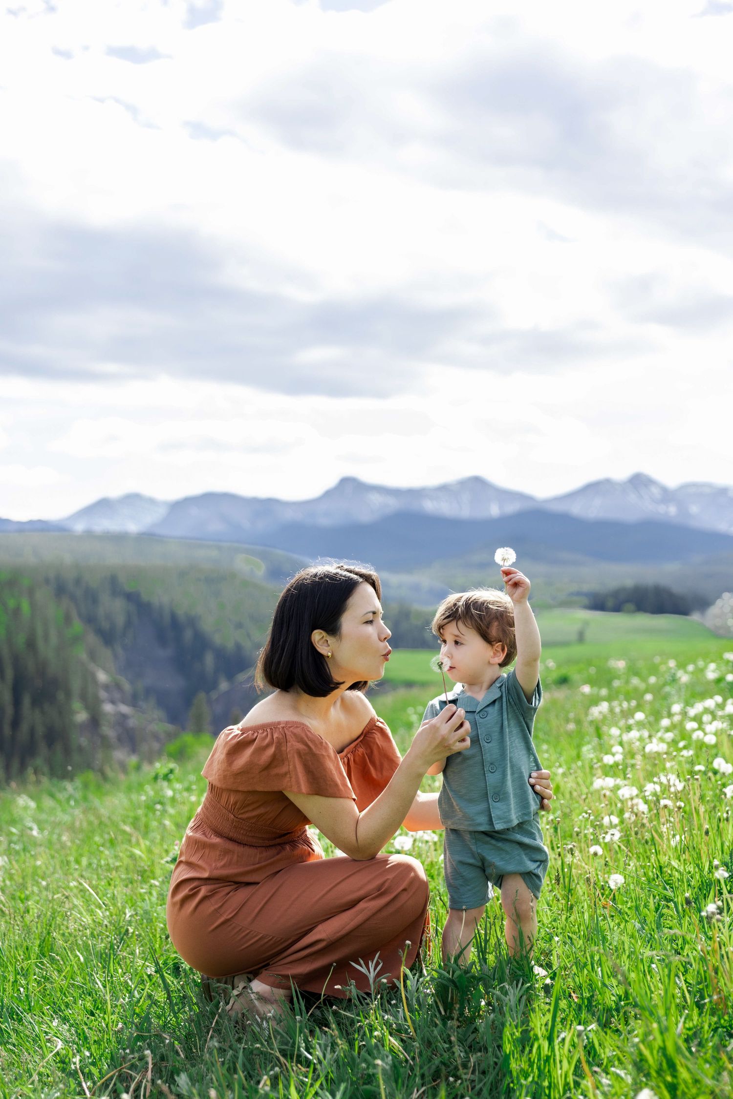 Mother and toddler blowing dandelions in a green meadow with mountain views on a cloudy day.