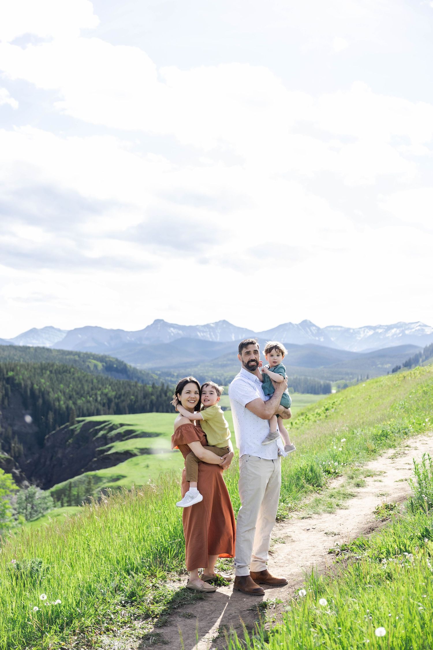 Family of four posing on a mountain trail with green meadows and snow-capped peaks in the background.